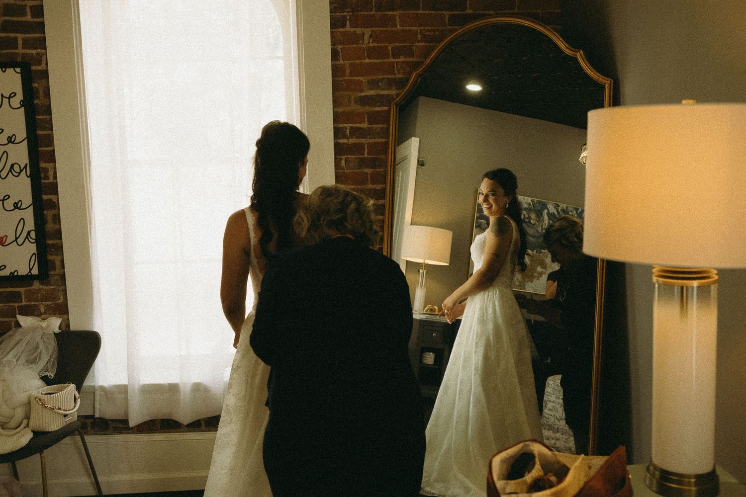 A bride in a white wedding dress stands in a room with exposed brick walls and table lamps.