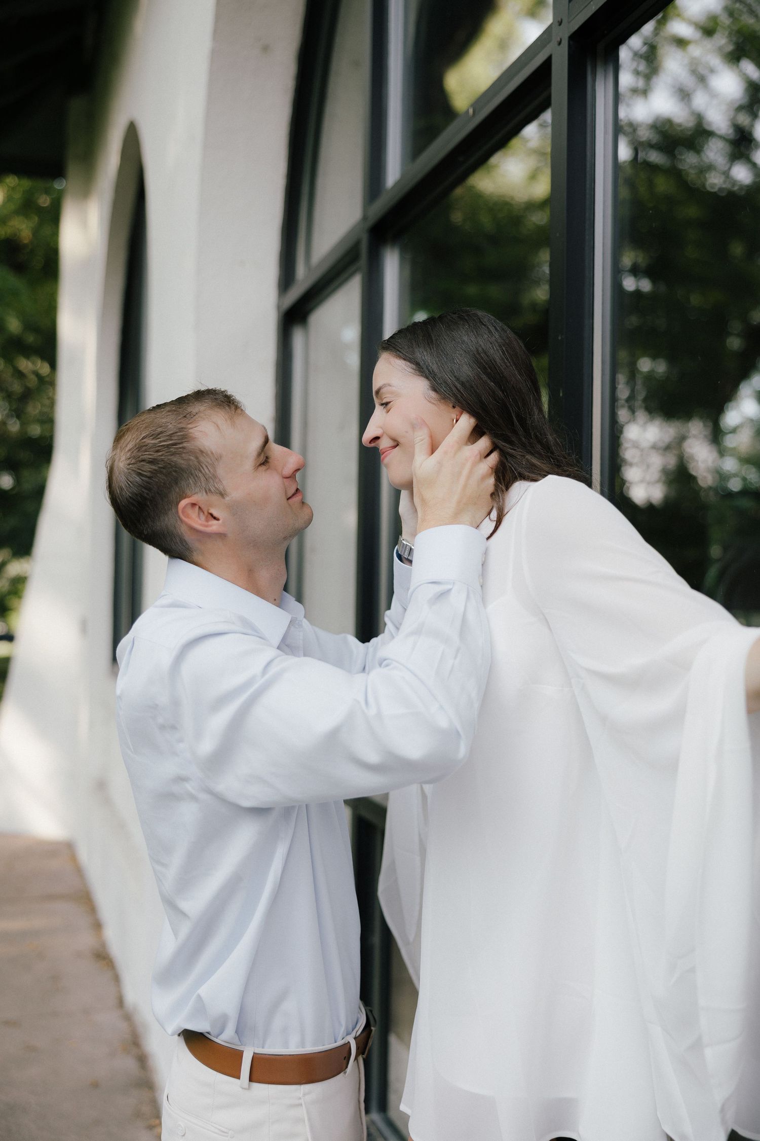 Romantic couple in matching white outfits sharing a tender moment outdoors near a building with large windows.