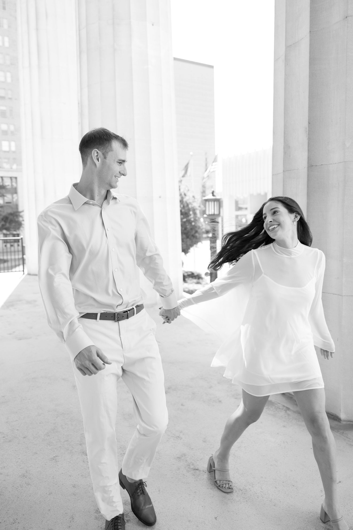 A joyful couple holds hands and walks between grand columns in a black and white engagement photo.