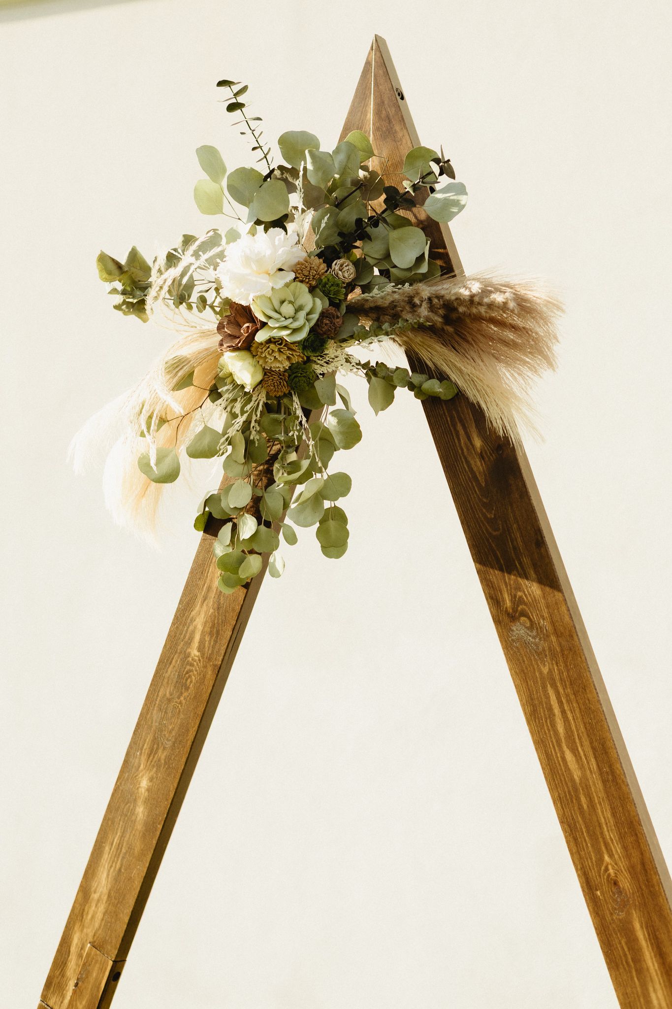Wooden triangular wedding arch decorated with eucalyptus leaves and floral arrangements.