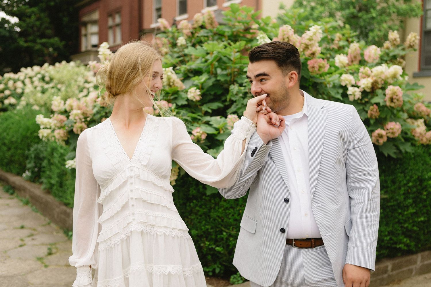 Couple in elegant attire sharing a romantic moment outdoors, man kissing woman's hand near blooming flowers.