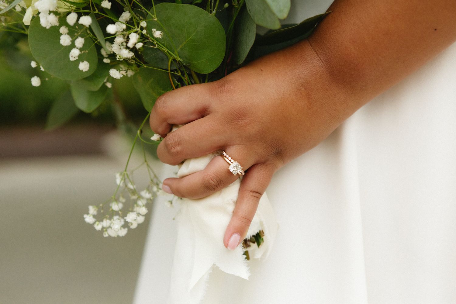 Close-up of hands holding white wedding bouquet with baby's breath flowers and elegant rings against white dress.