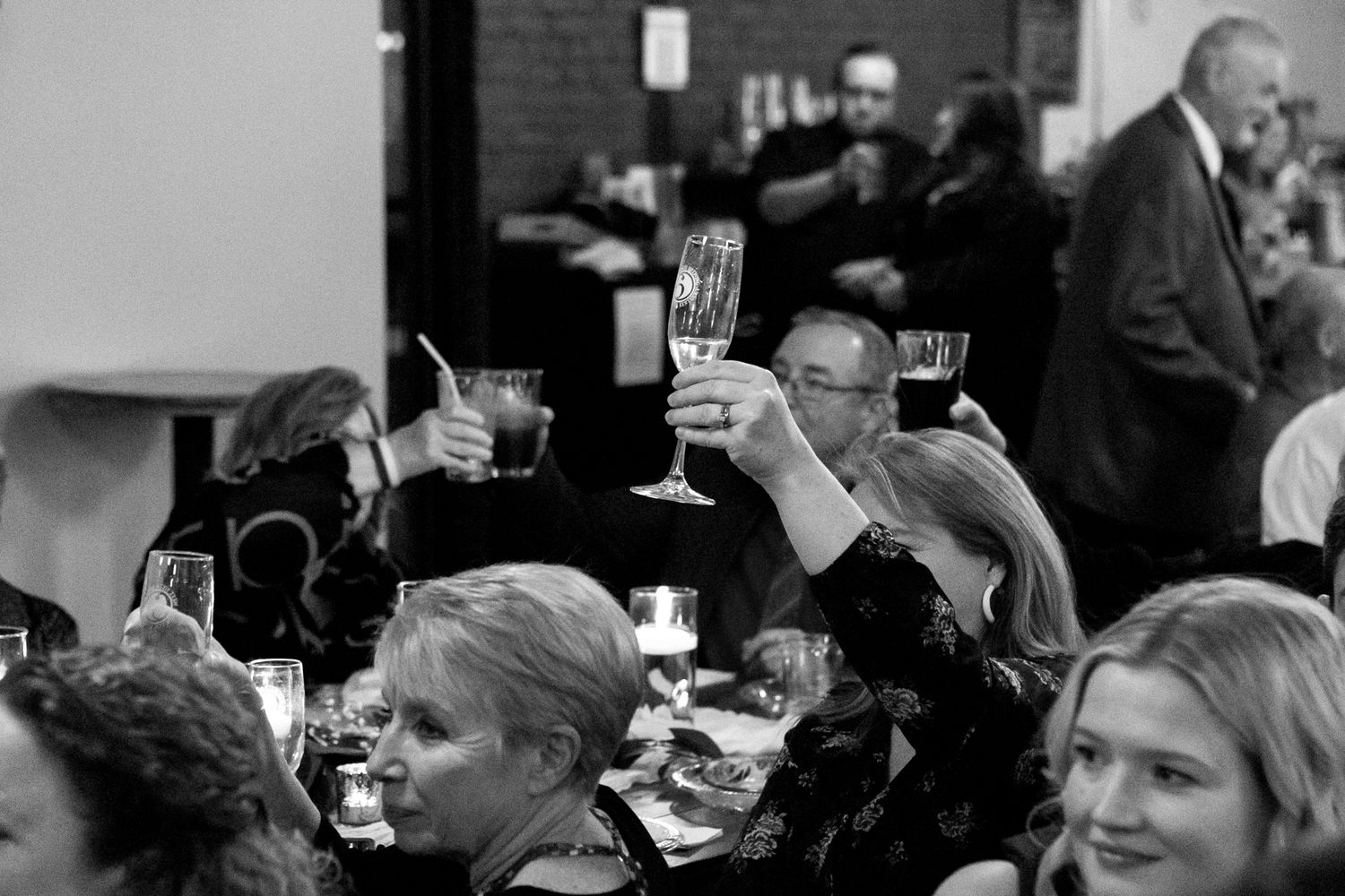 Wedding guests raise glasses in celebration during a black and white reception scene.