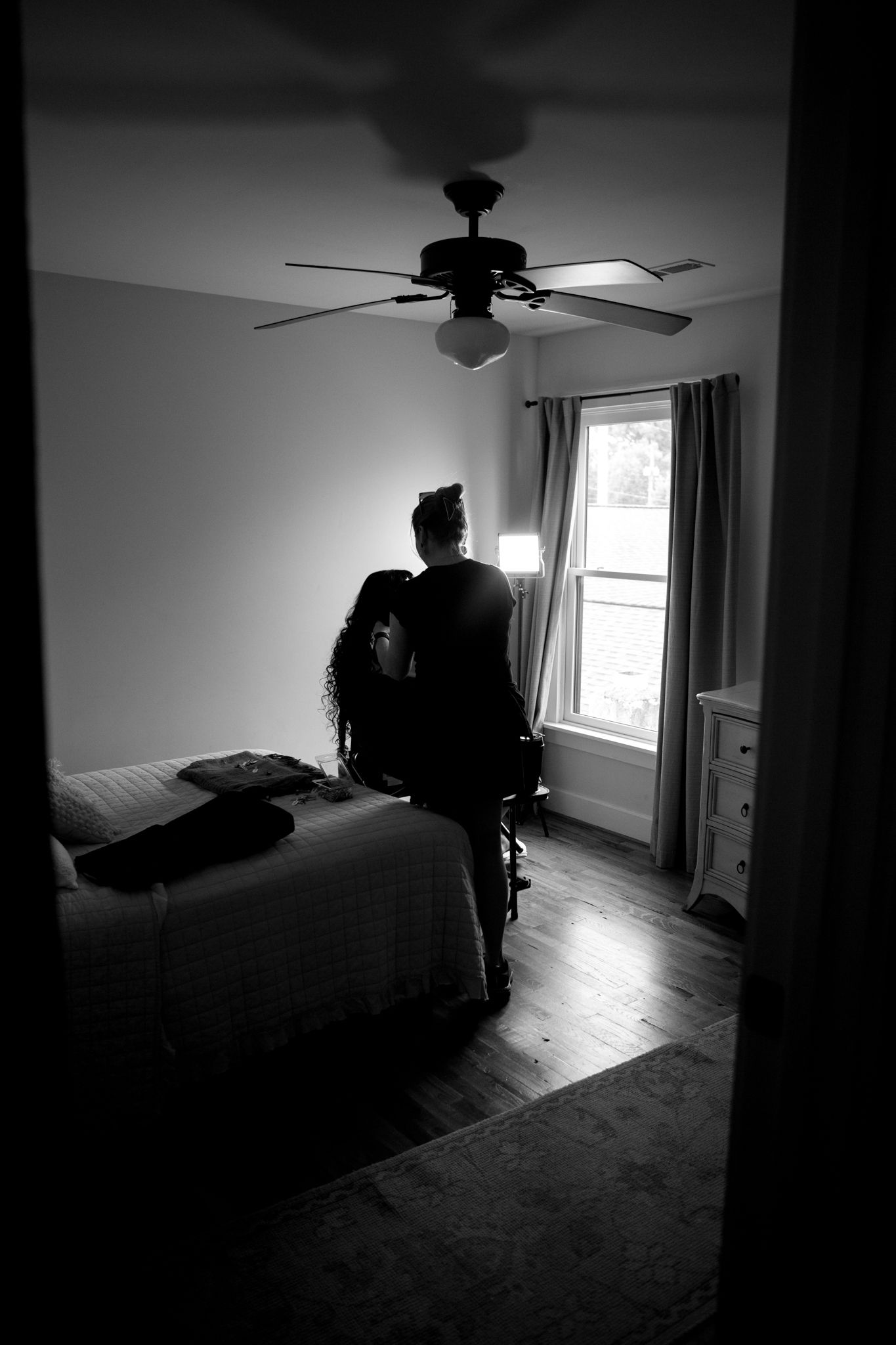 A silhouetted figure stands in dark bedroom with ceiling fan and window, creating dramatic black and white contrast.