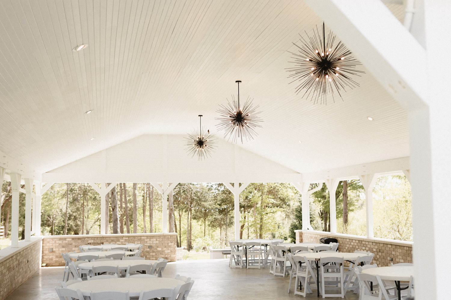 A bright white wedding reception pavilion with starburst chandeliers and round tables set up for an outdoor celebration.