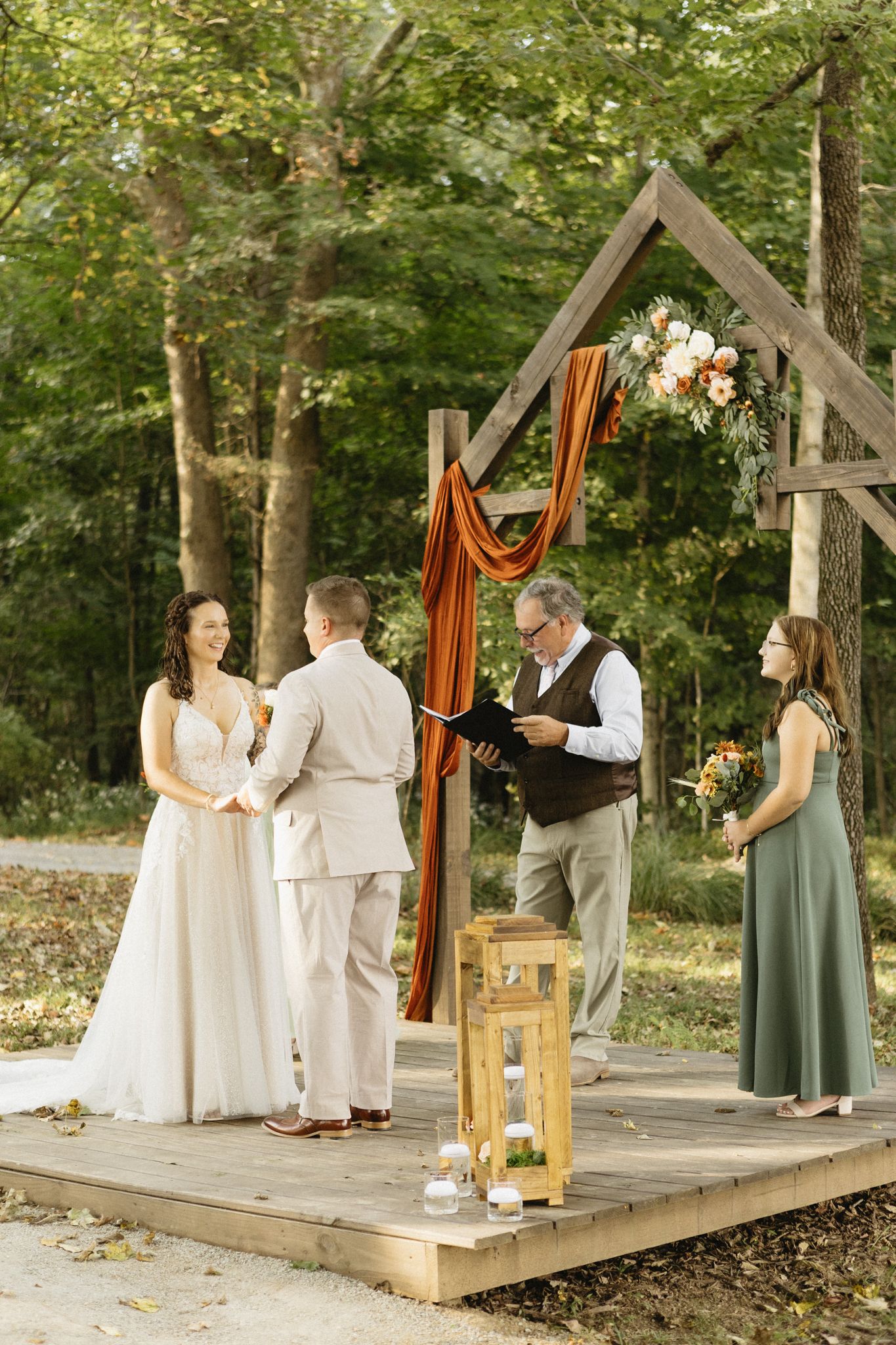 Outdoor wedding ceremony on a wooden platform with orange draping and natural forest backdrop.