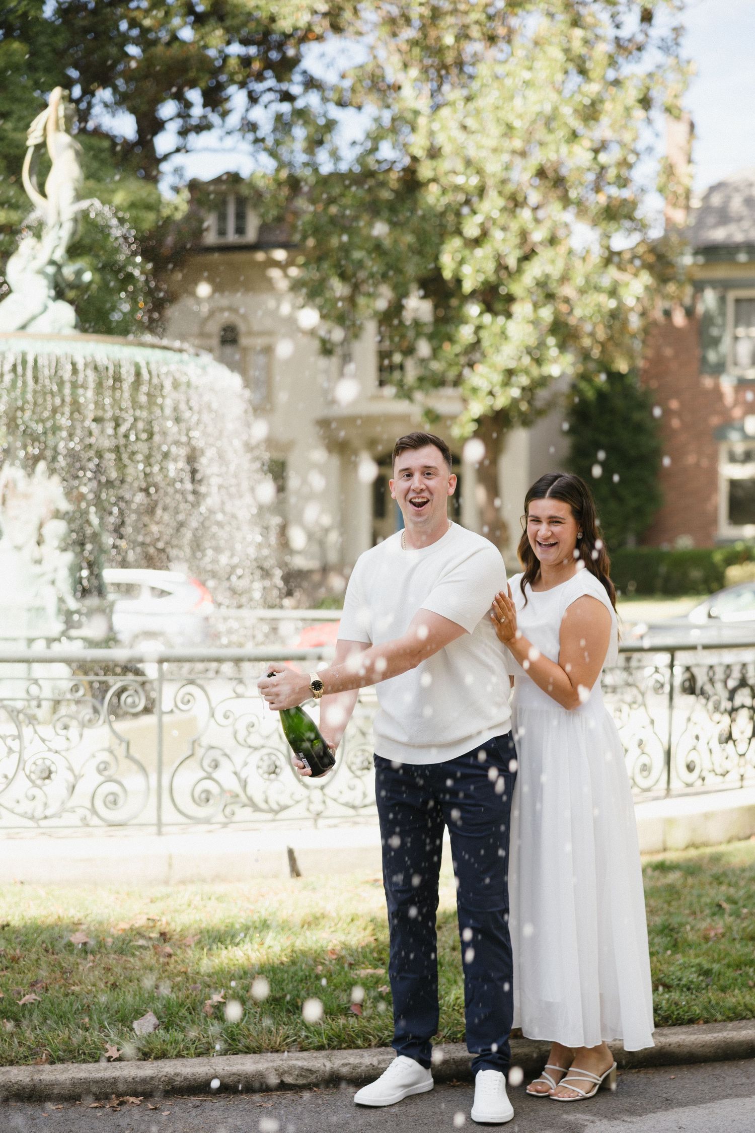 Joyful couple popping champagne bottle by ornate fountain, woman in white dress celebrating outdoors.