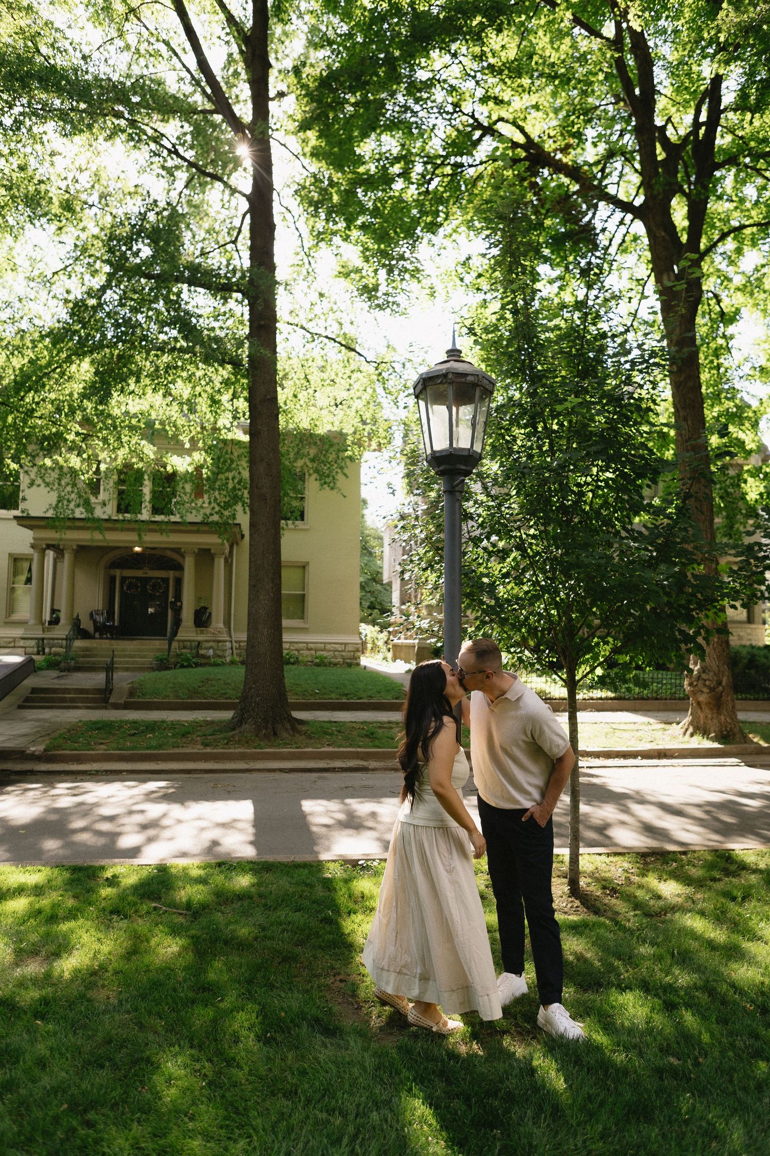 Couple sharing a romantic moment by a vintage lamppost surrounded by lush green trees near a historic building.