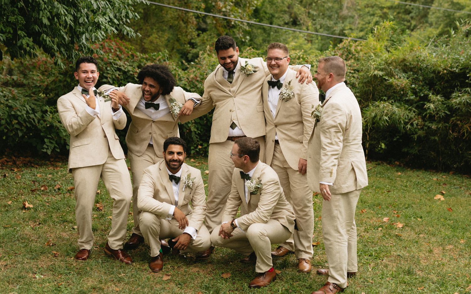 Wedding party wearing beige and cream colored suits poses playfully on grass with trees in background.