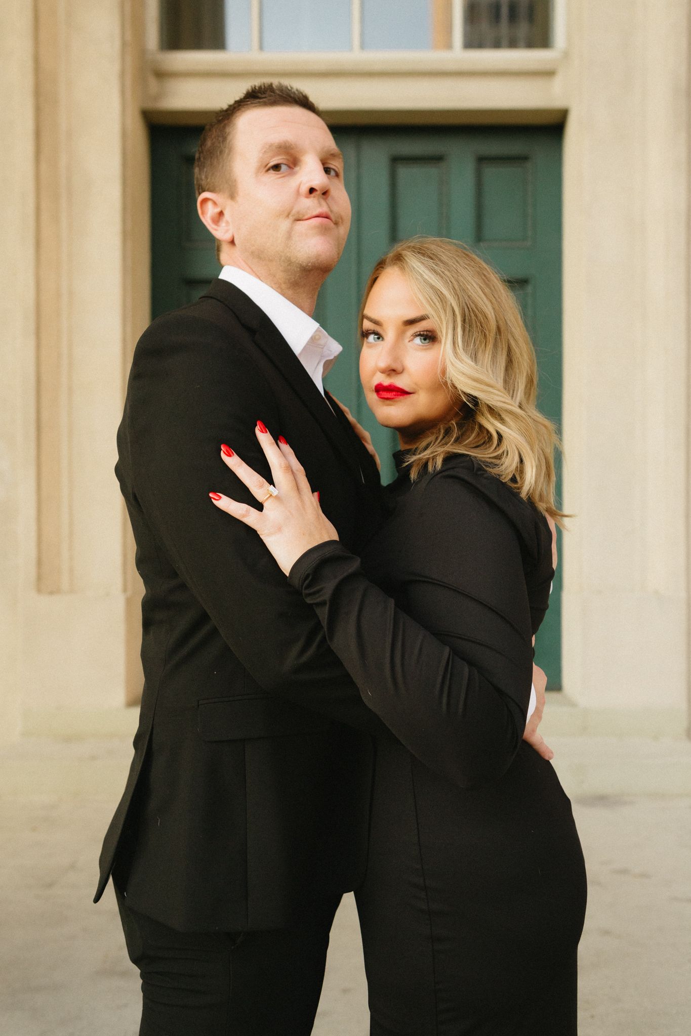 A couple in matching black formal attire poses elegantly against a light-colored building with a green door.