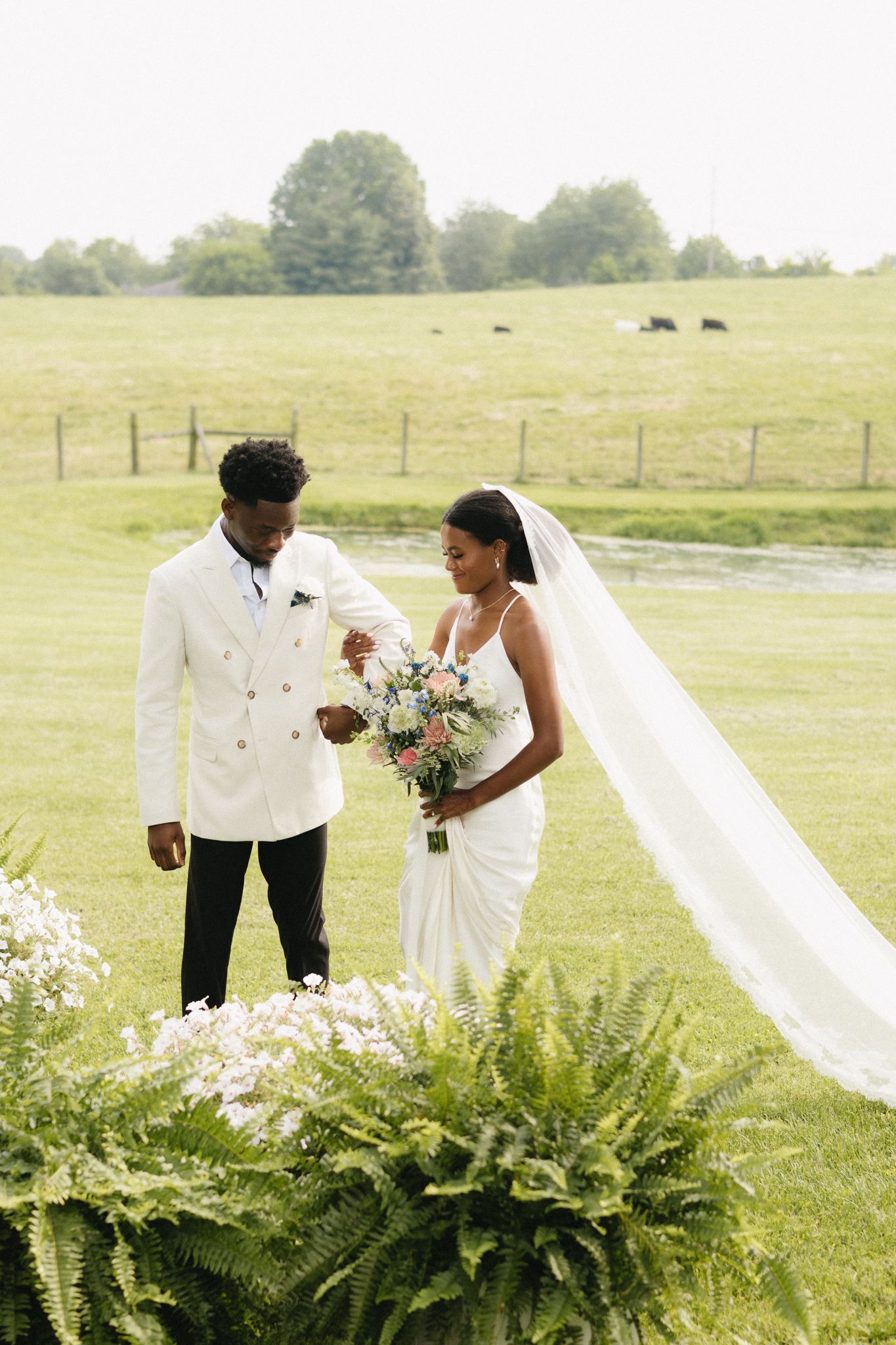A wedding couple stands in a meadow with ferns and wildflowers as the bride holds a beautiful bouquet.