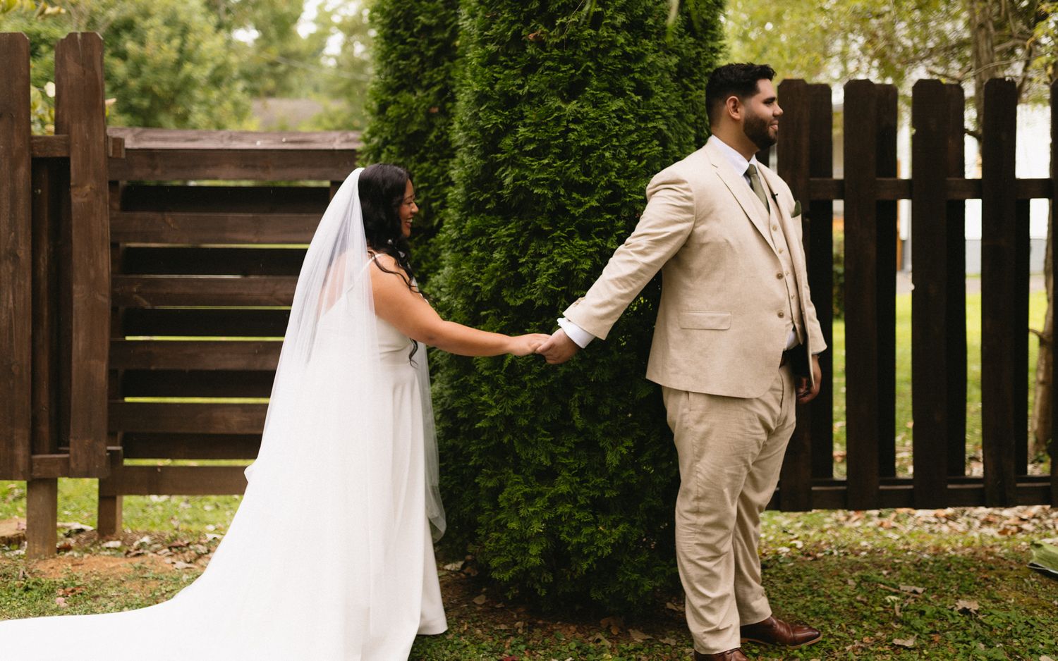A bride and groom hold hands while standing at opposite sides of a rustic wooden fence corner during their outdoor wedding.