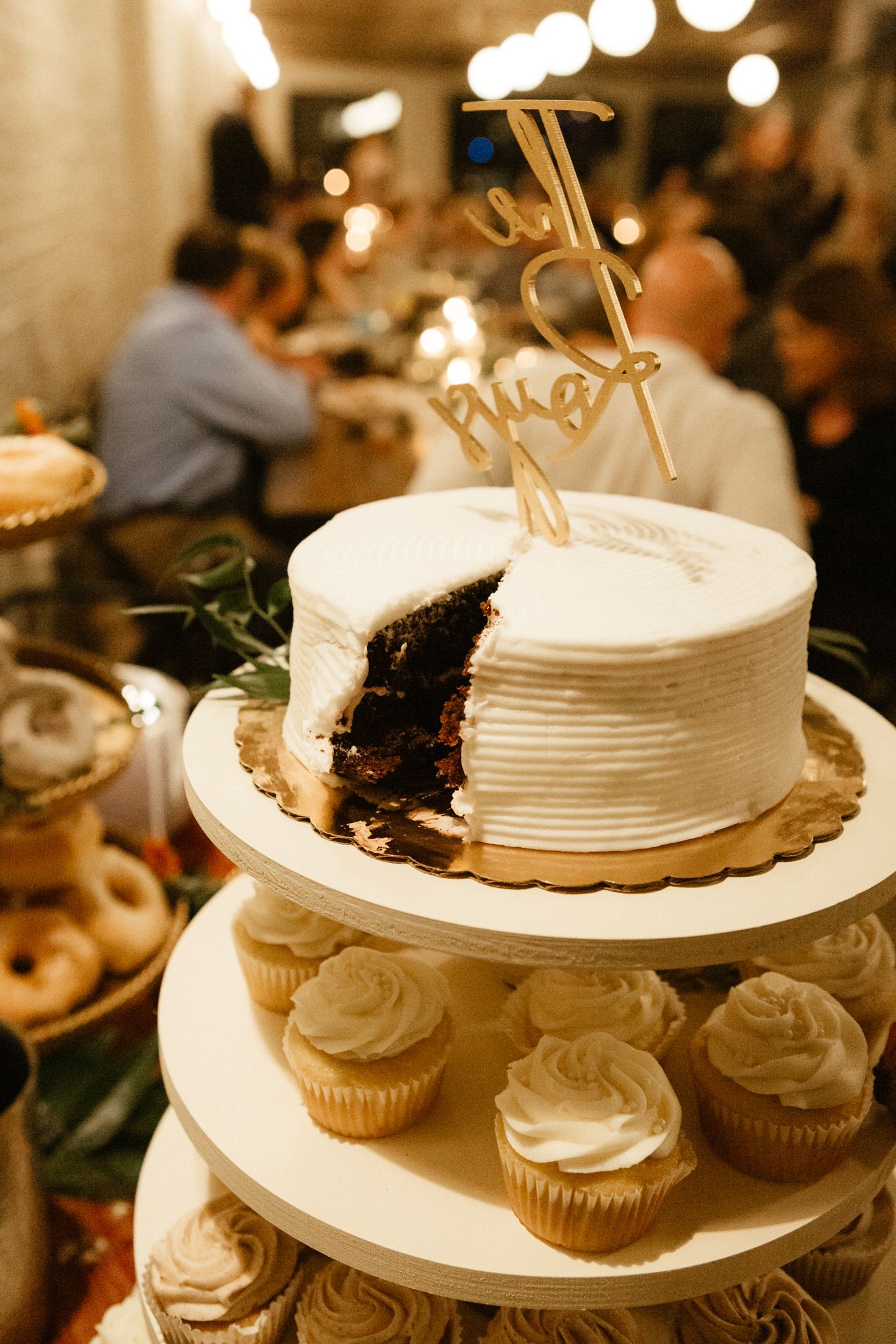 A white wedding cake with ribbed frosting sits on a tiered stand surrounded by cupcakes in warm lighting.