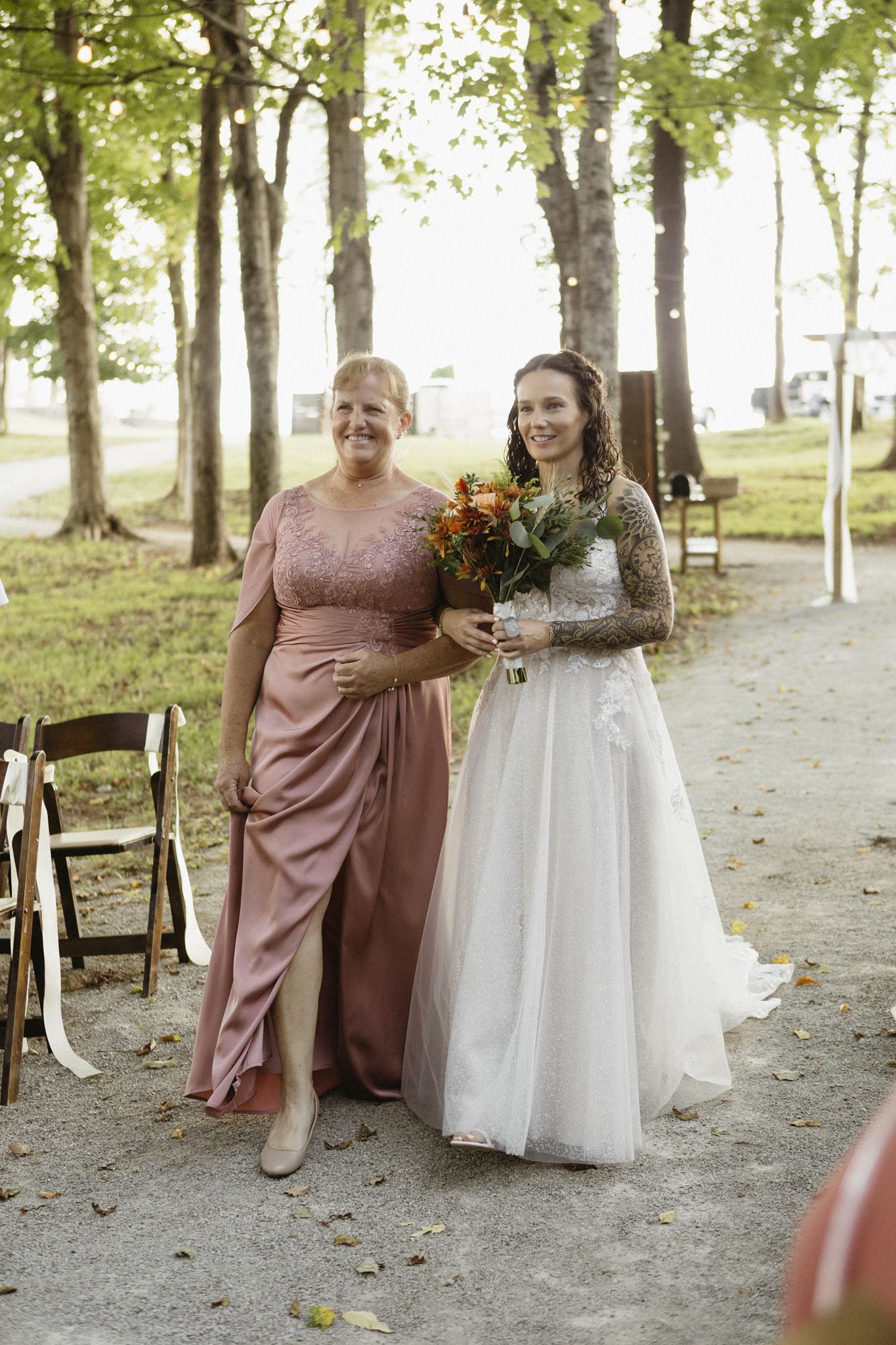 Two women in formal dresses stand together on a tree-lined path during an outdoor wedding celebration.