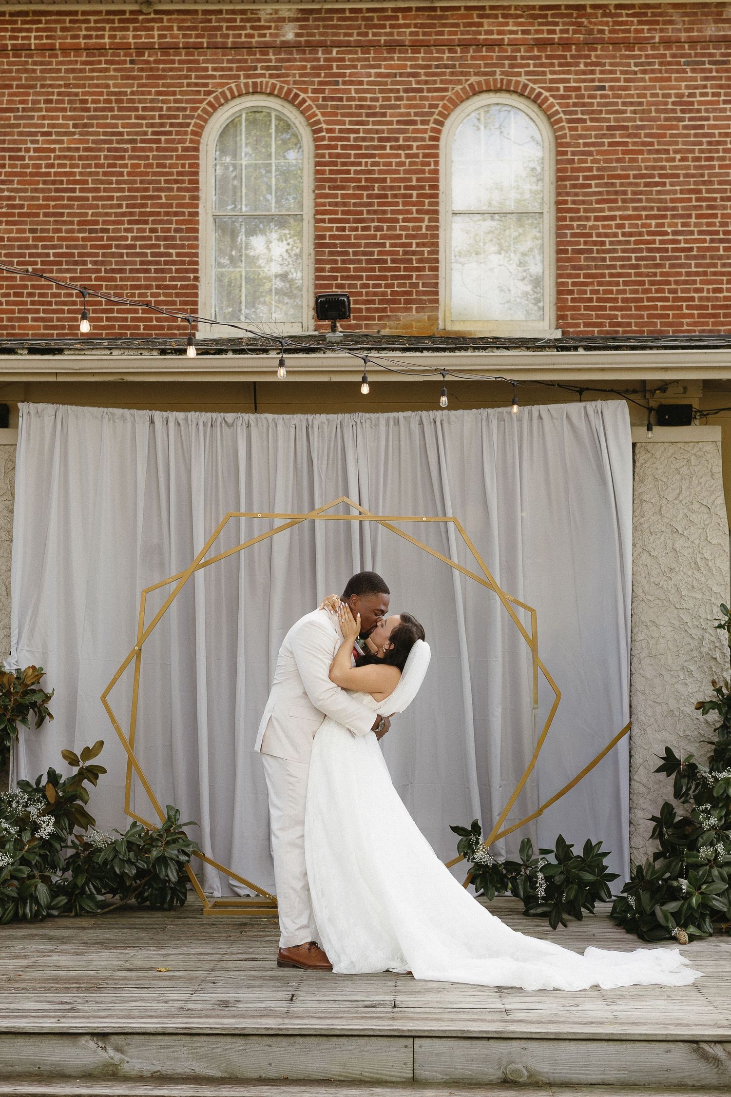 Wedding couple embracing under a geometric arch with white drapes against a brick wall.