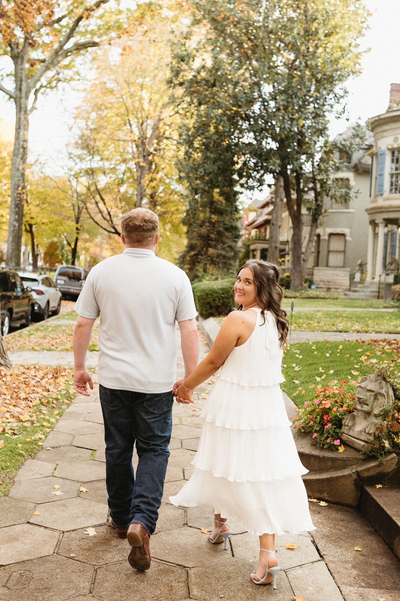 A couple walks hand in hand down a stone pathway lined with autumn trees and Victorian houses.