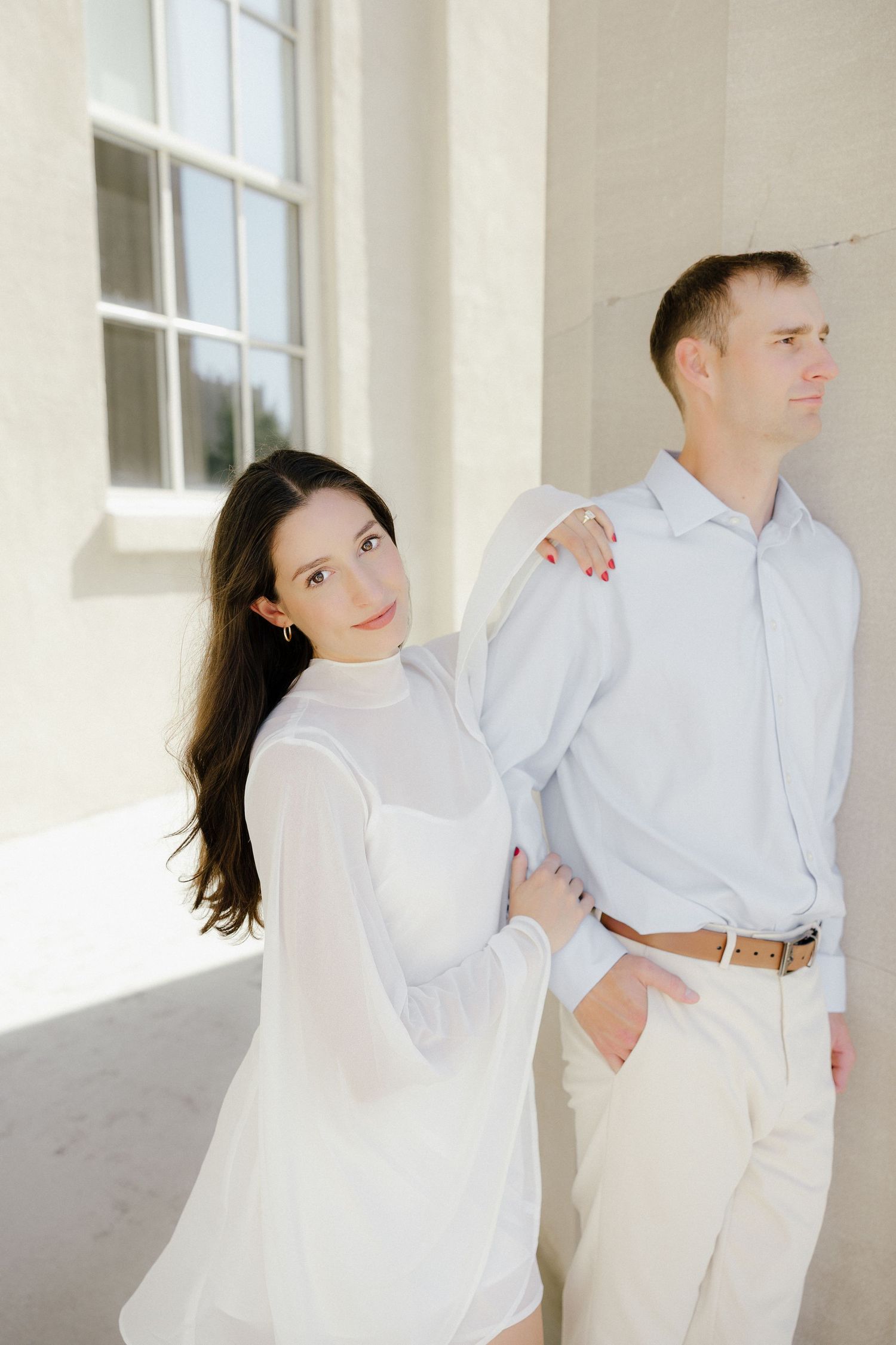 Woman in white dress embracing man from behind against a bright white wall in soft natural light.