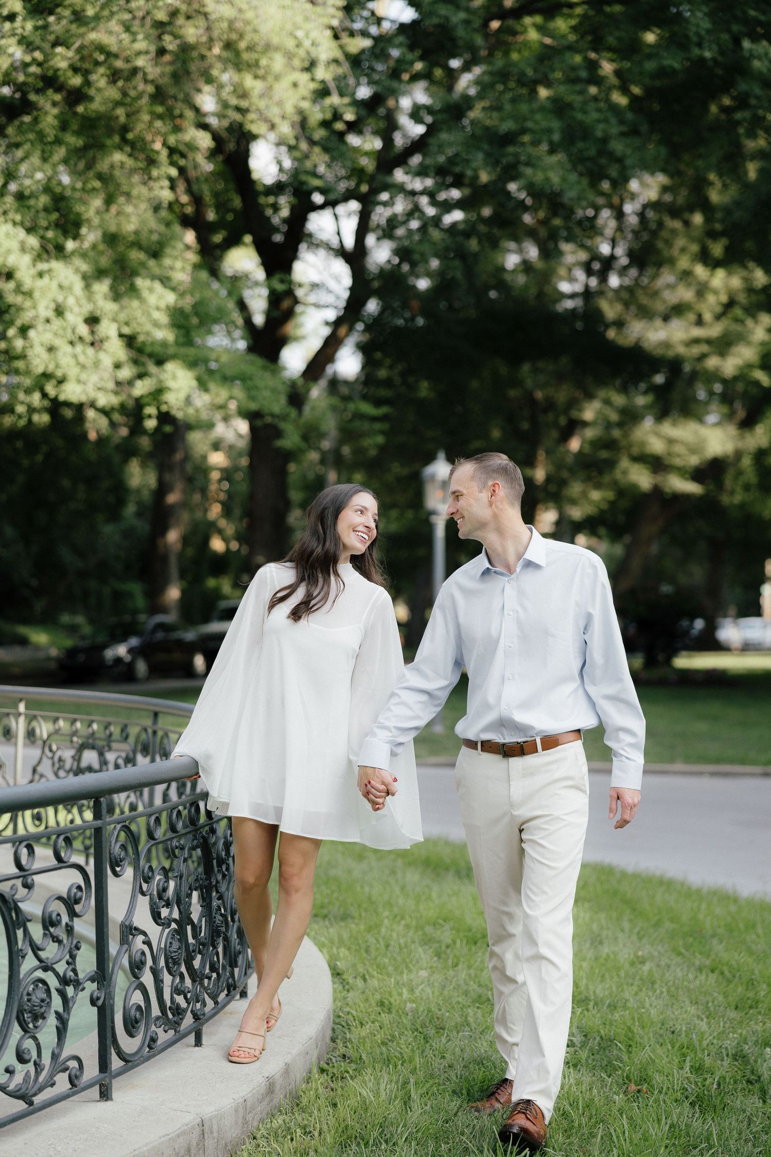 Happy couple in white outfits holding hands and smiling while walking near ornate iron railing in park.
