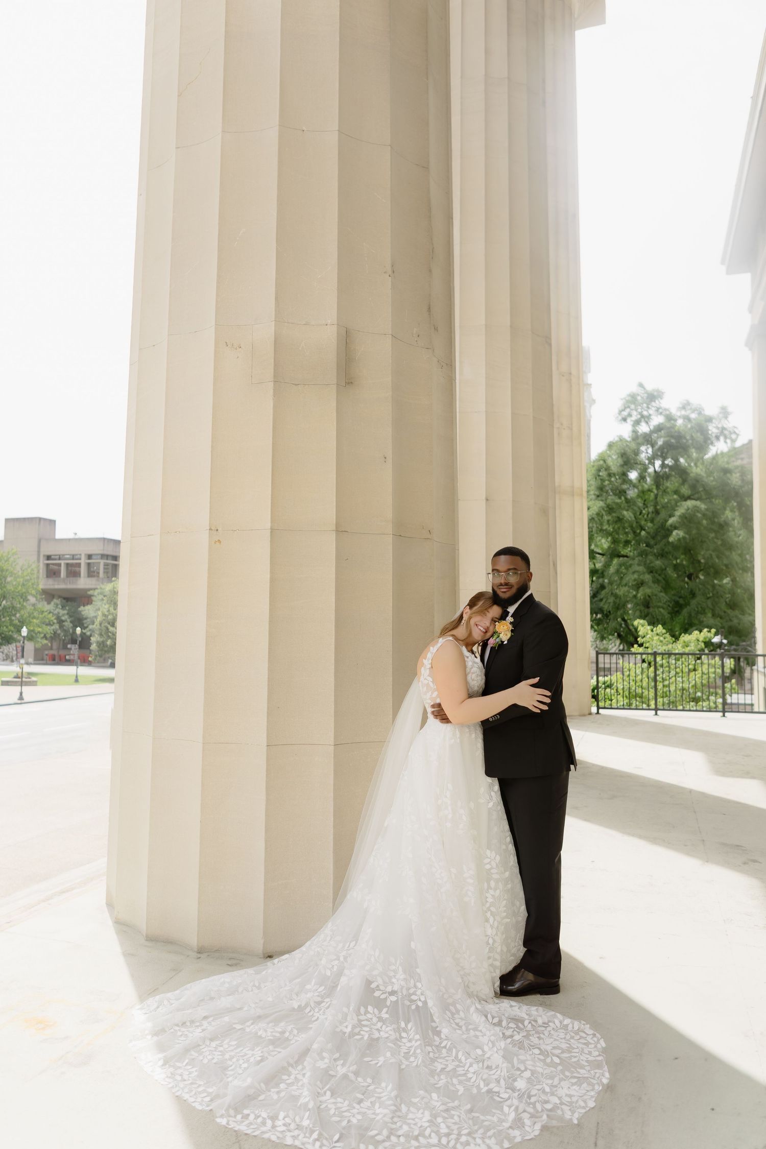 Bride and groom embrace between grand white columns, her lace cathedral veil trailing elegantly on marble steps.