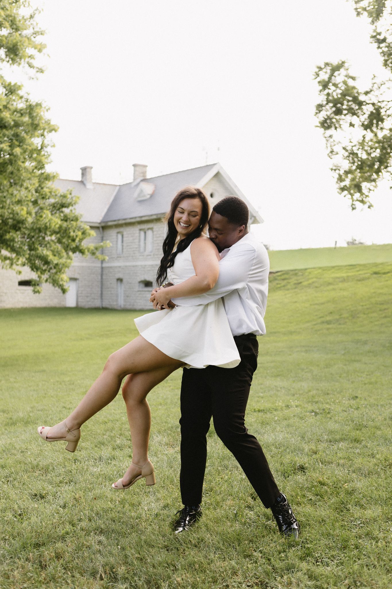 A joyful couple dances on green grass, man lifting smiling woman in white dress, elegant stone mansion in background.