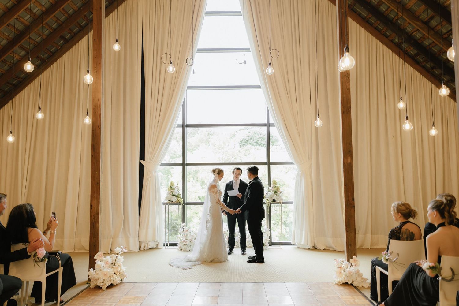 Bride and groom stand at altar in rustic barn venue with white drapes and string lights.