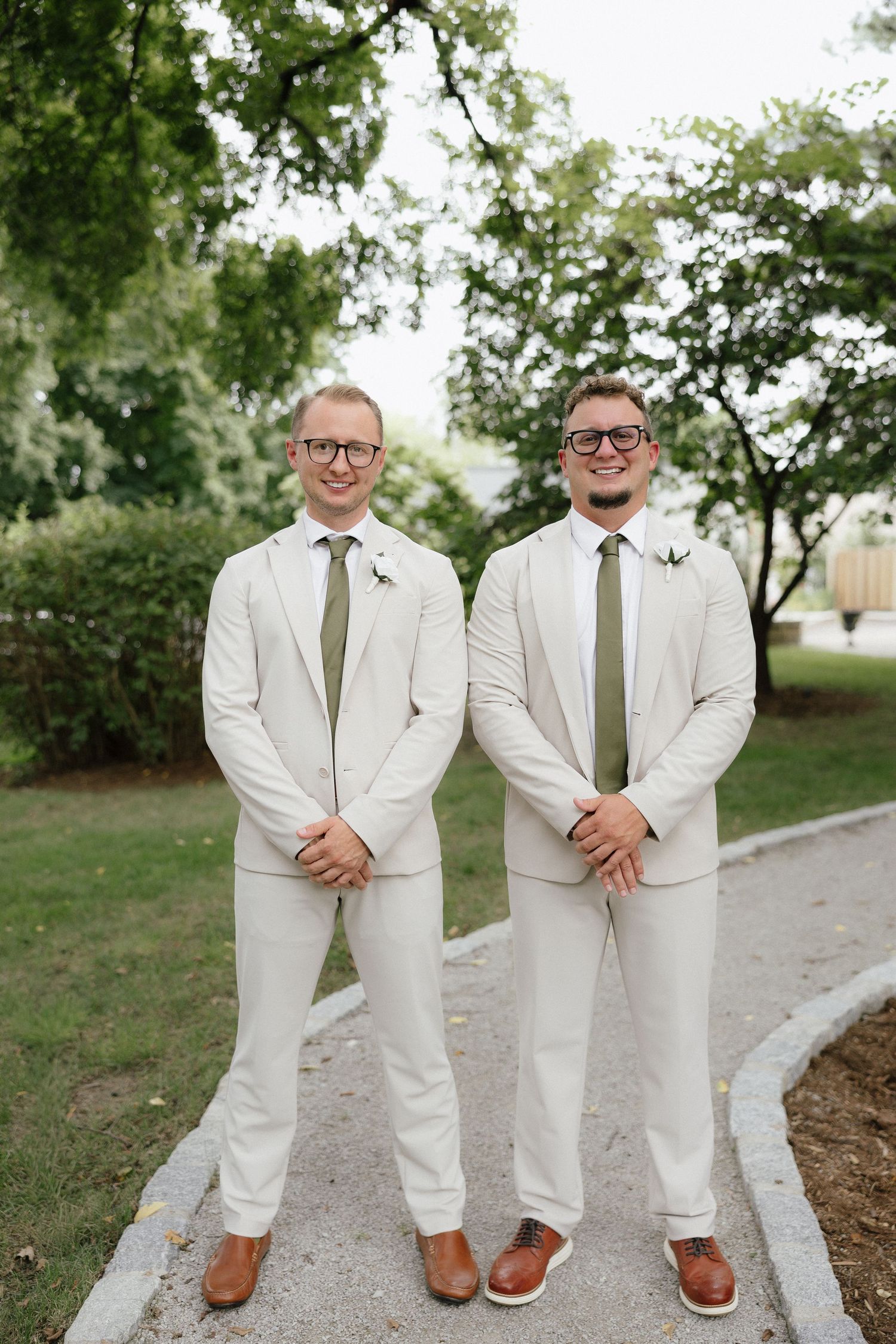 Two groomsmen in matching cream suits with olive green ties and glasses smile outdoors at wedding.