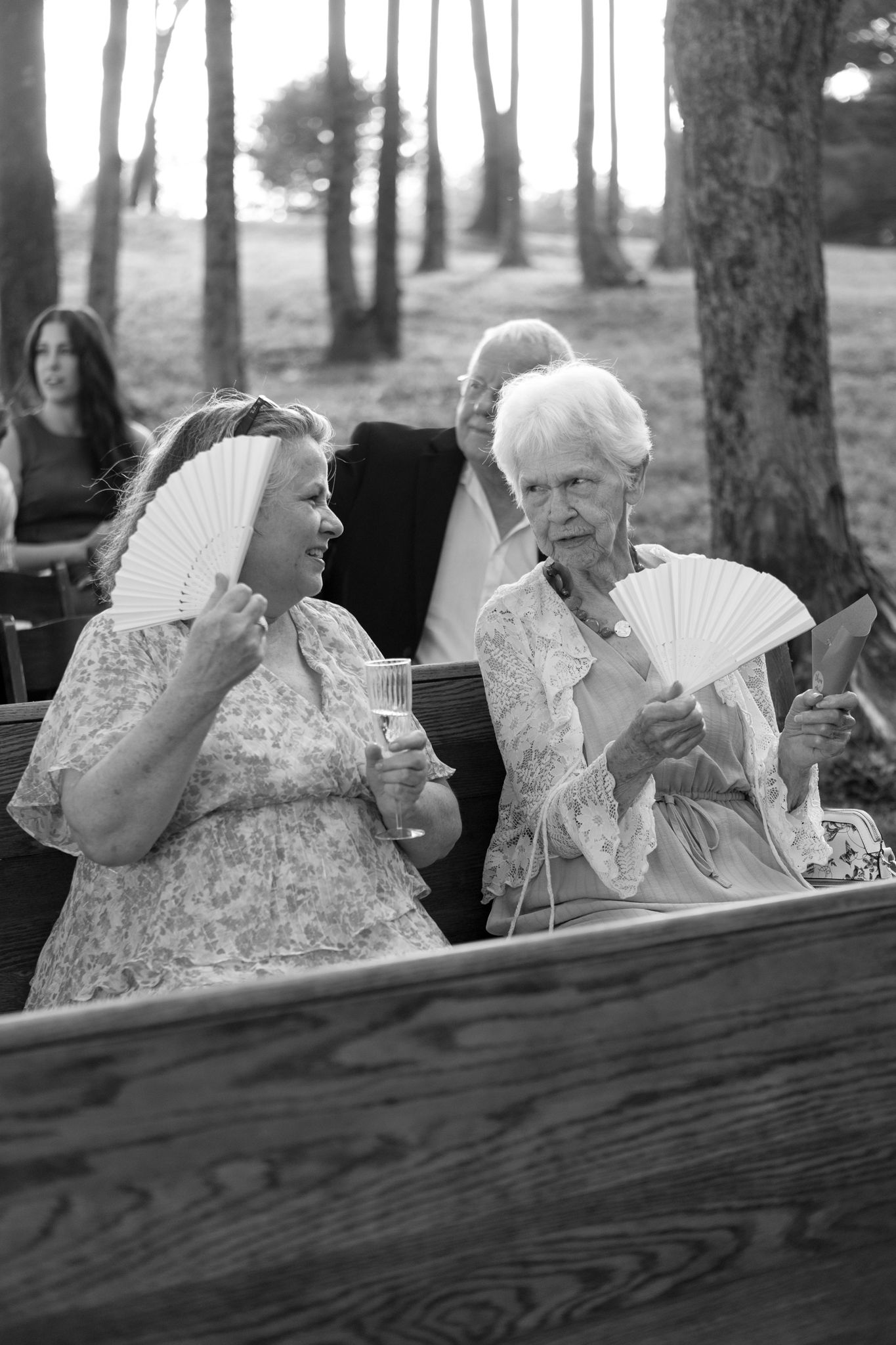 A black and white photo of people sitting on benches in a wooded area holding paper fans on a summer day.