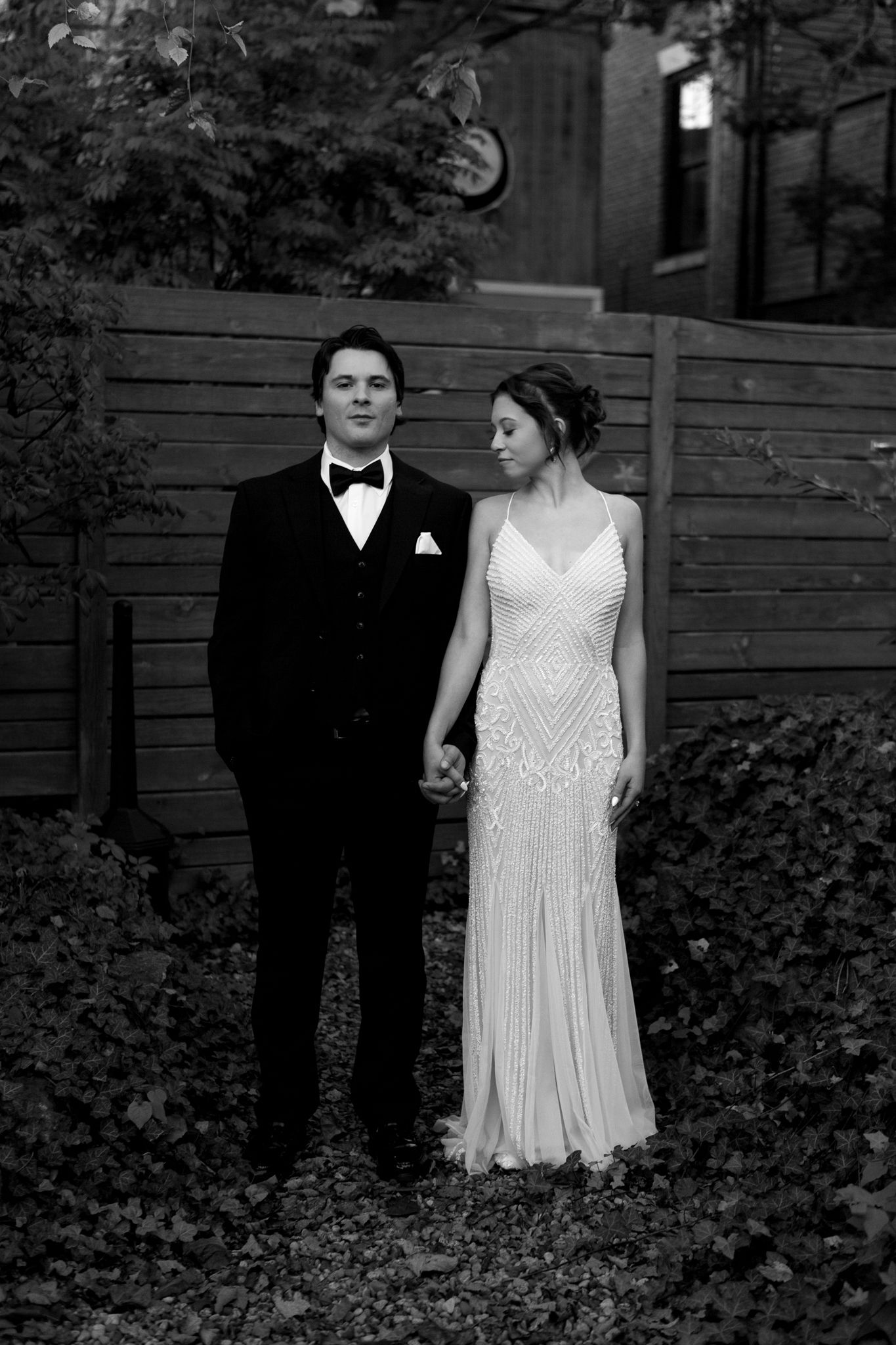 A couple in formal attire stands together in a garden setting wearing a white dress and black tuxedo.