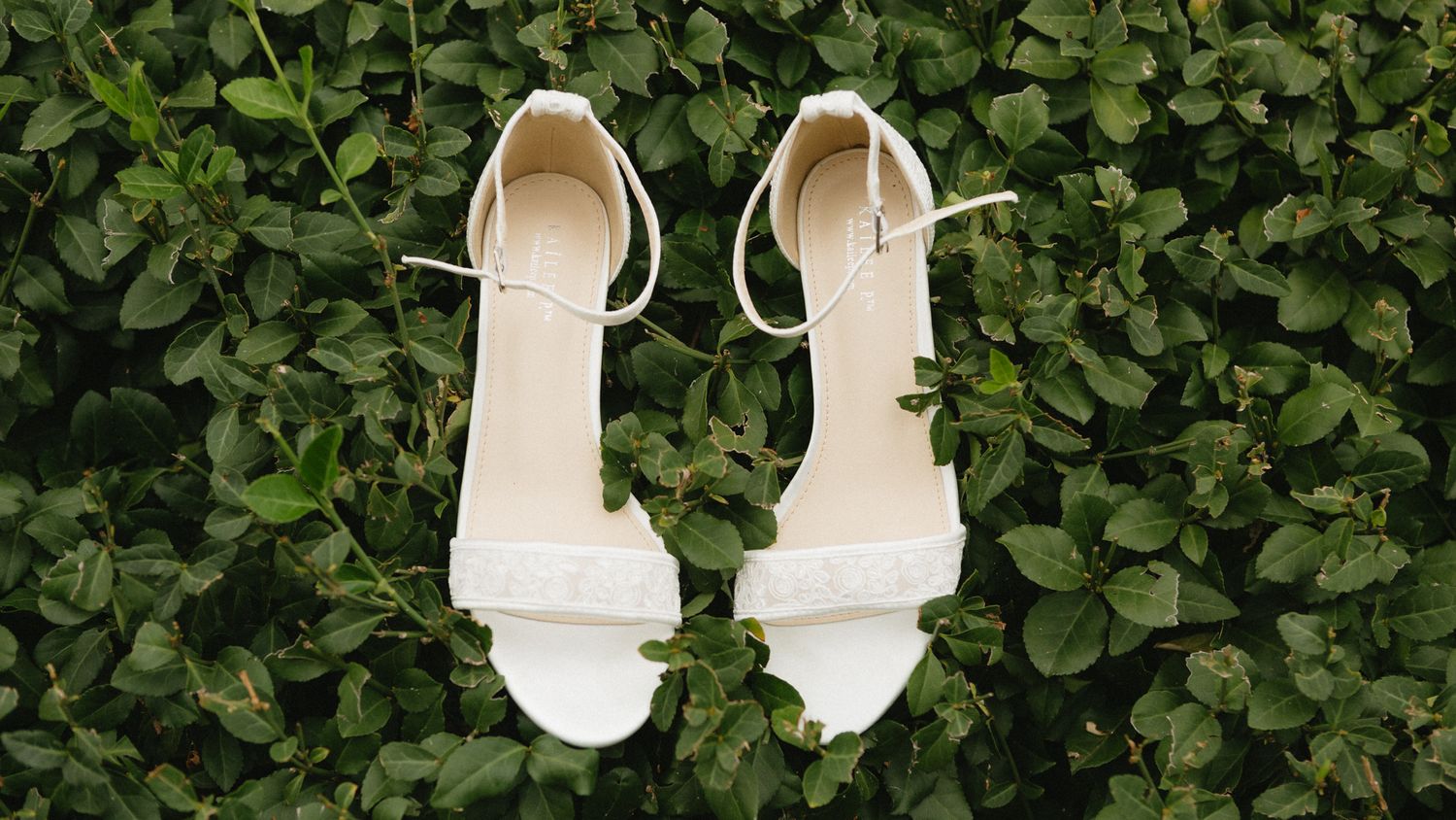 Elegant white bridal sandals with ankle straps laid on green leafy background.