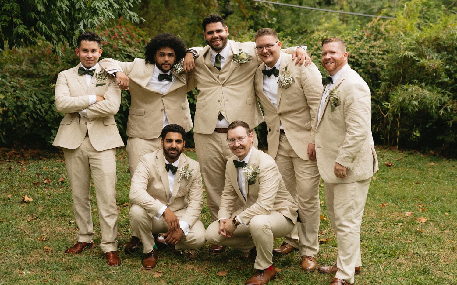 A wedding party in matching beige suits poses together outdoors on a lawn with greenery in the background.