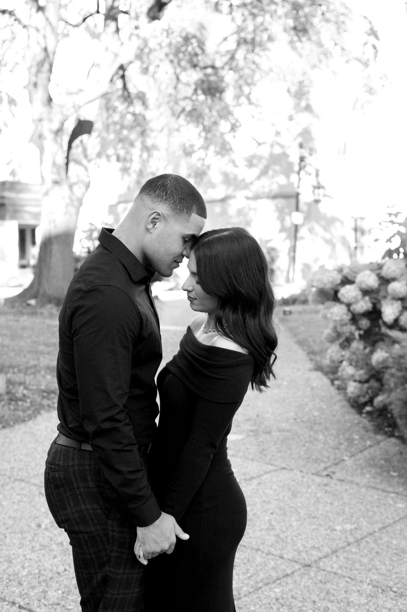 A tender black and white photo of a couple in matching dark outfits sharing an intimate moment in a garden.