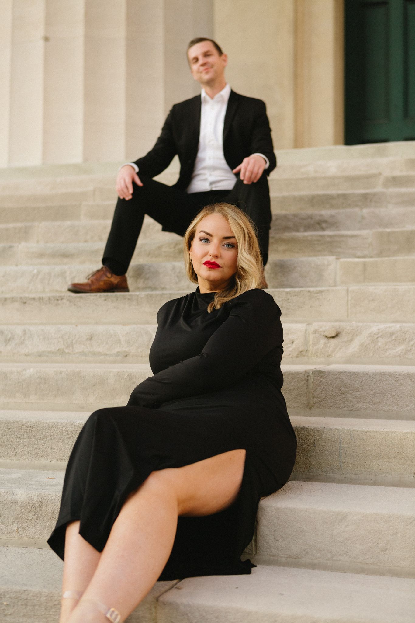 A stylish couple poses on grand stone steps wearing formal black attire for an elegant photoshoot.