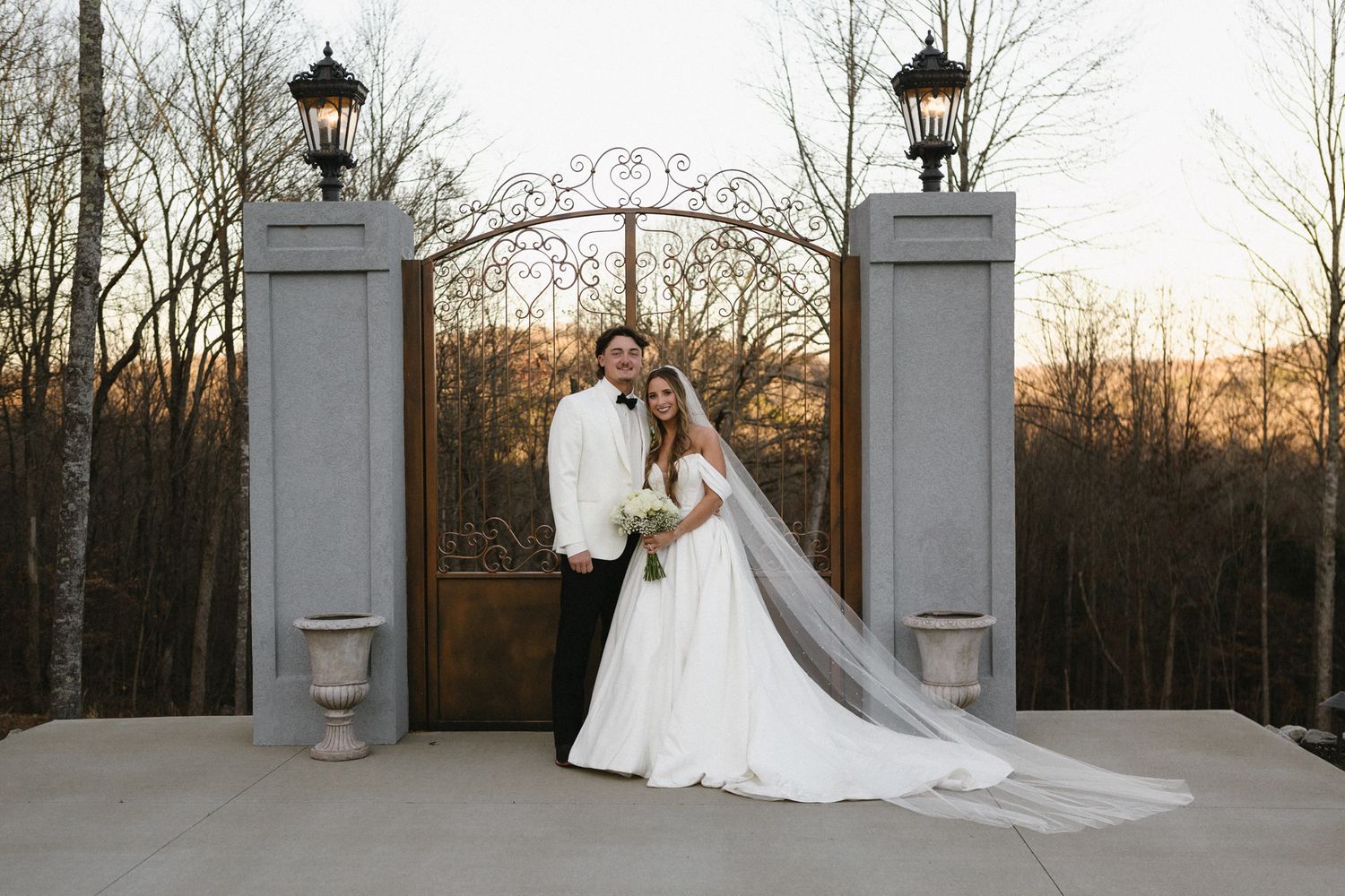 A stunning bride in a flowing white gown stands elegantly by ornate stone pillars at sunset.