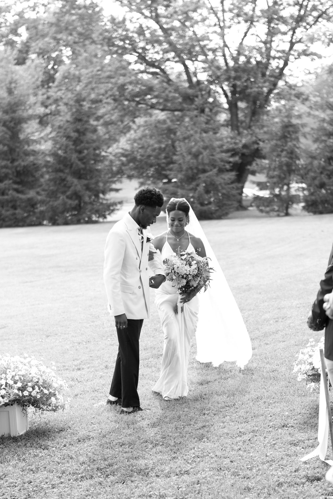 A black and white photo captures a tender moment between a bride and groom on a grassy lawn surrounded by trees.
