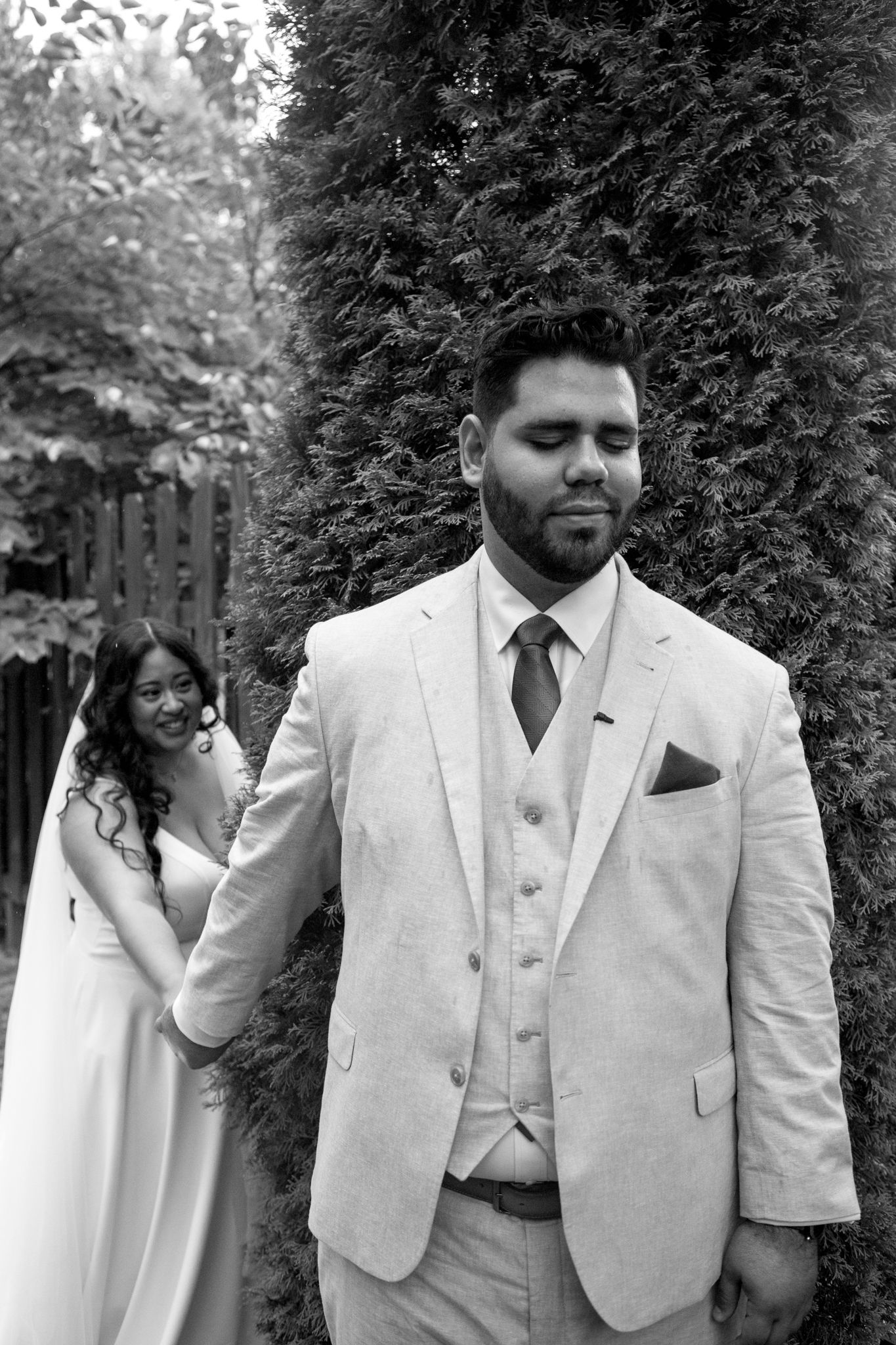Black and white wedding photo shows bride in dress reaching for groom in light colored suit with vest and tie by garden wall.
