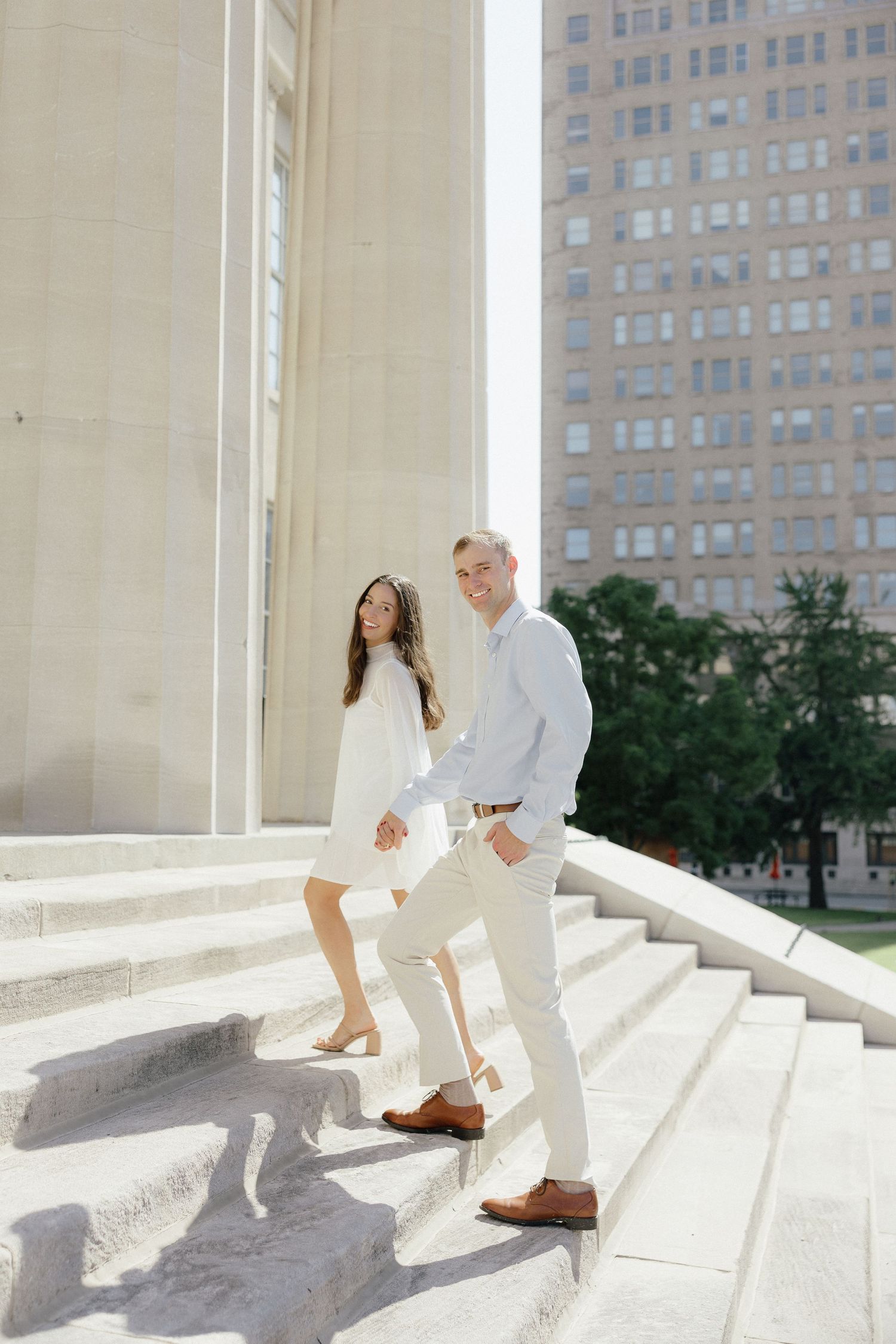 Couple in white outfits posing on marble steps near tall columns and a city building on a sunny day.