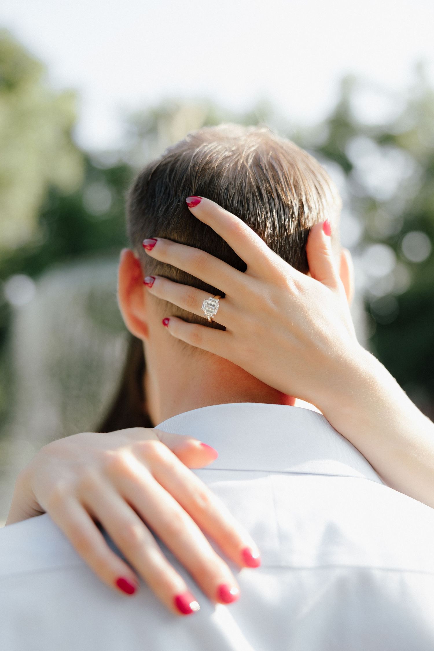 Woman showing diamond engagement ring with red nails while embracing a man outdoors.