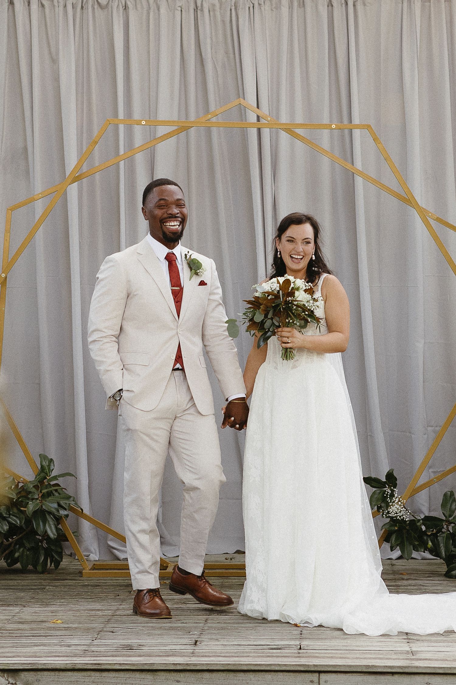 Wedding couple poses on a stage with geometric gold decorations against a grey curtain backdrop.