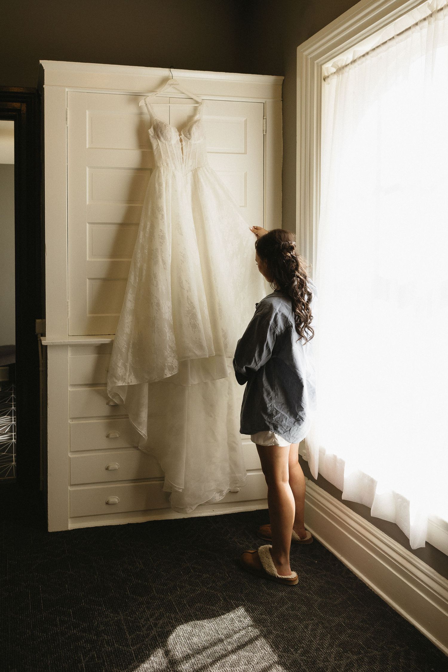 A wedding dress hangs on display while someone in casual attire looks out a nearby window.