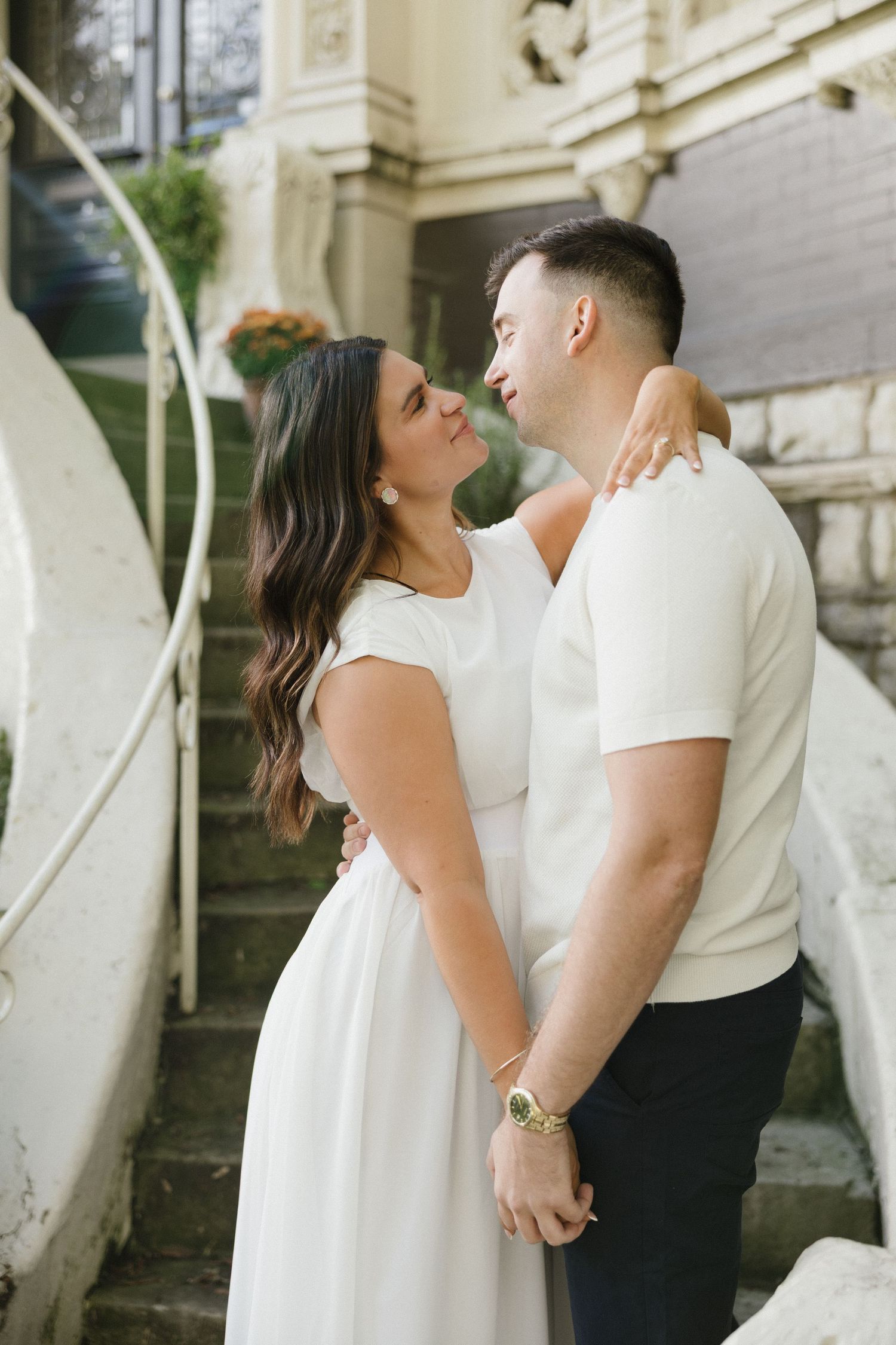 A couple shares a romantic moment near vintage stone steps, woman in white dress and man in cream shirt holding hands and almost kissing.