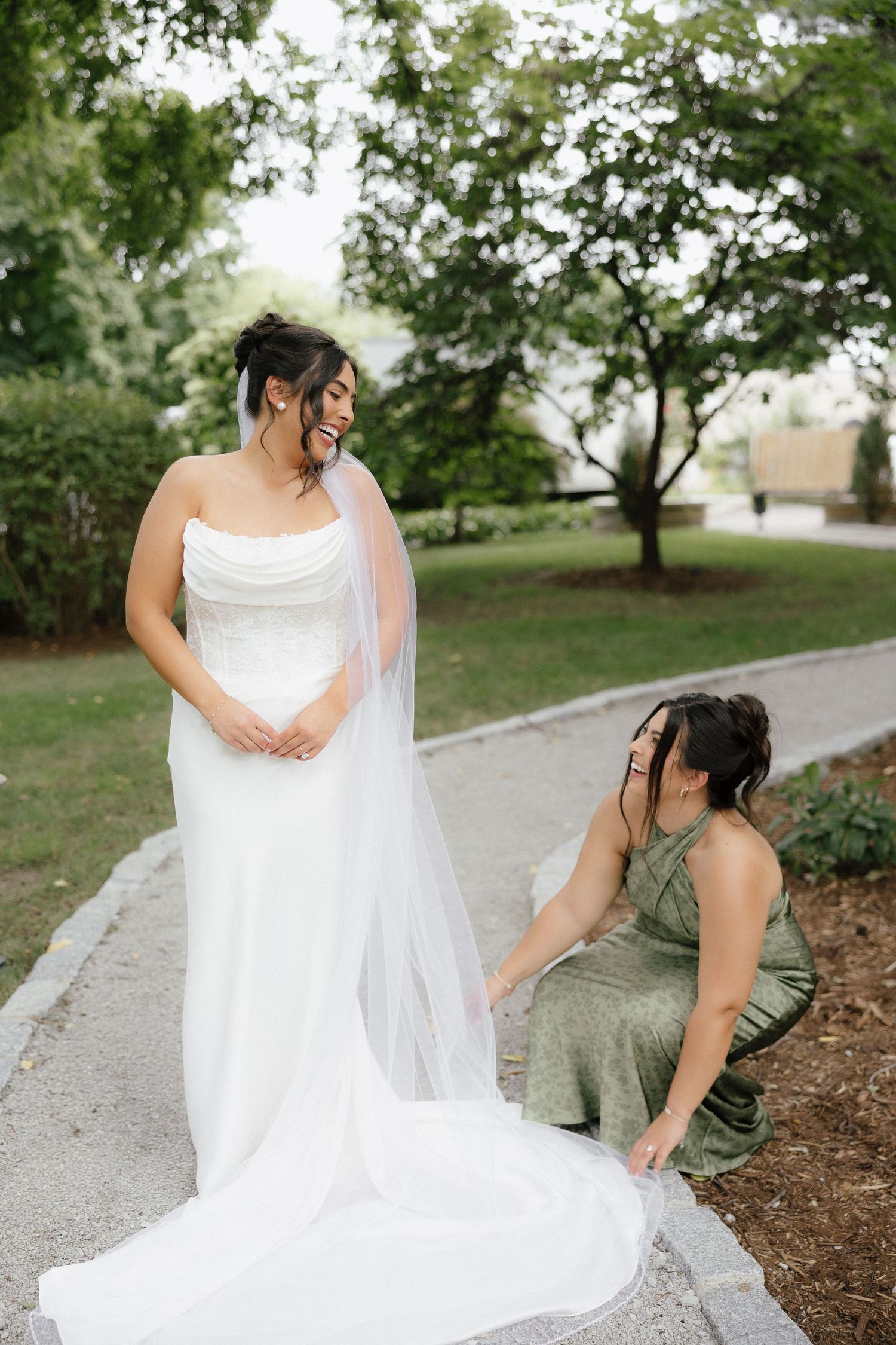 Bridesmaid in sage green dress adjusting bride's cathedral veil outdoors.