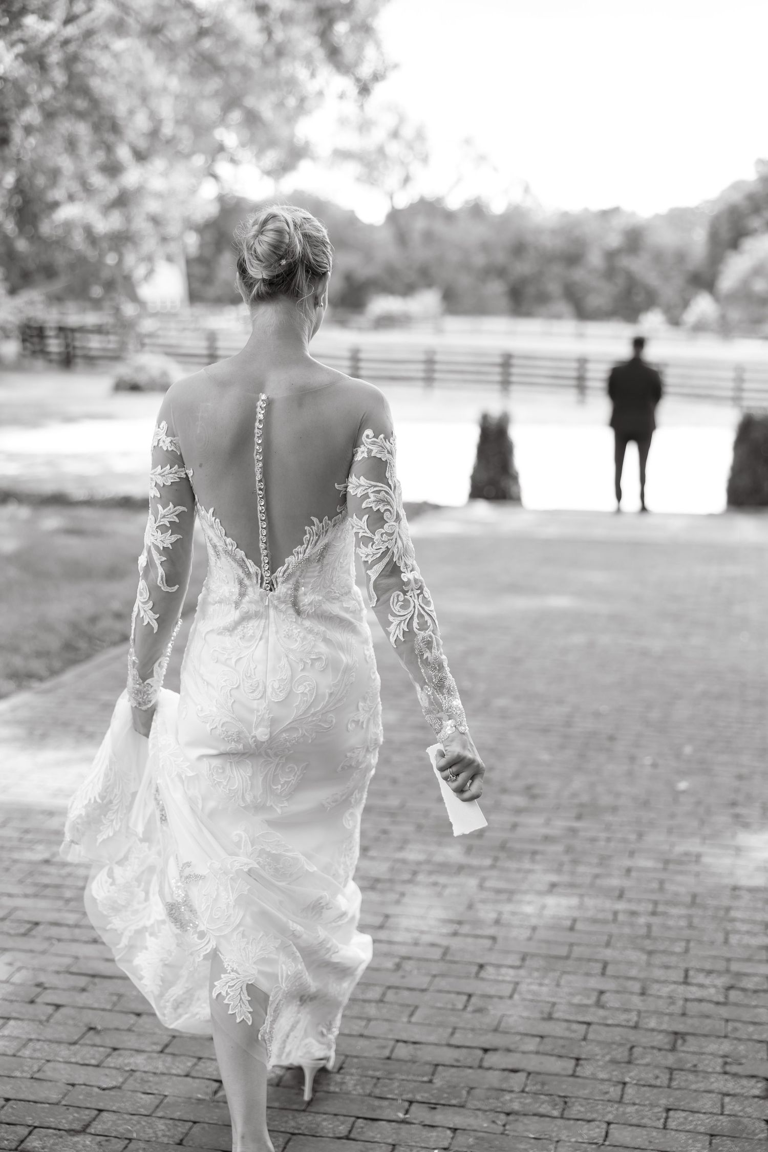 Bride in lace backless wedding gown walking toward groom outdoors in elegant black and white photo.