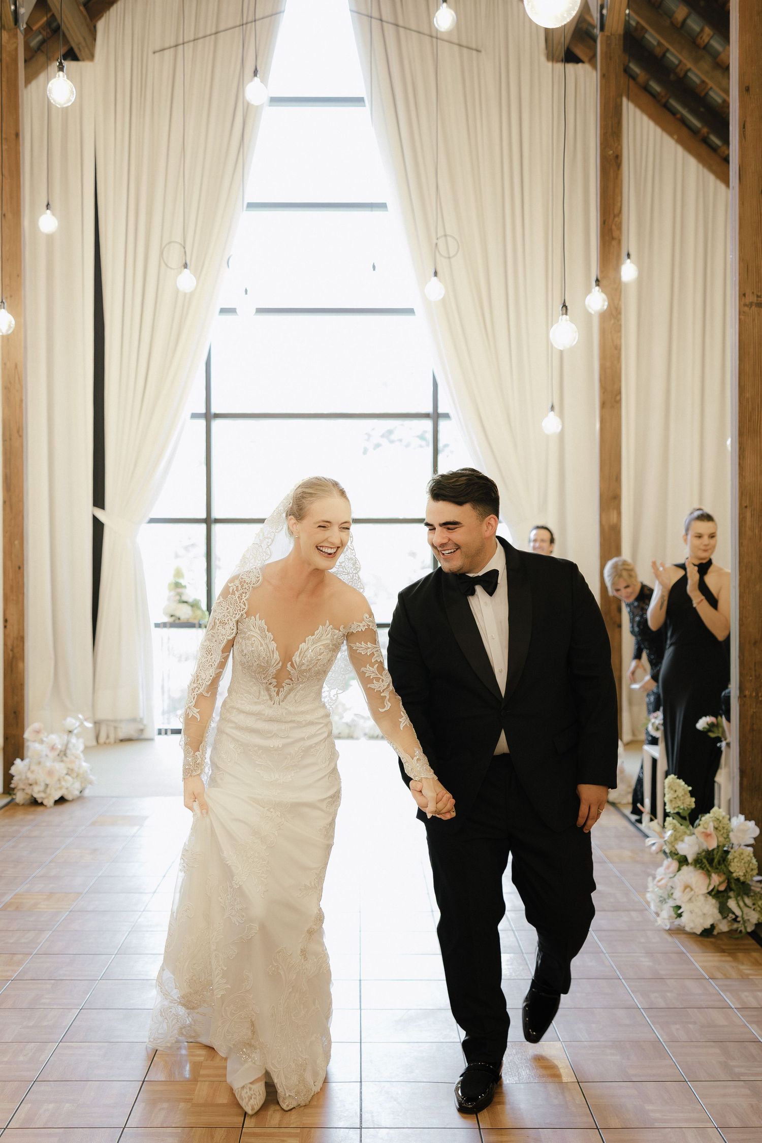 Bride in lace gown and groom in tuxedo walk hand in hand during their wedding reception entrance.