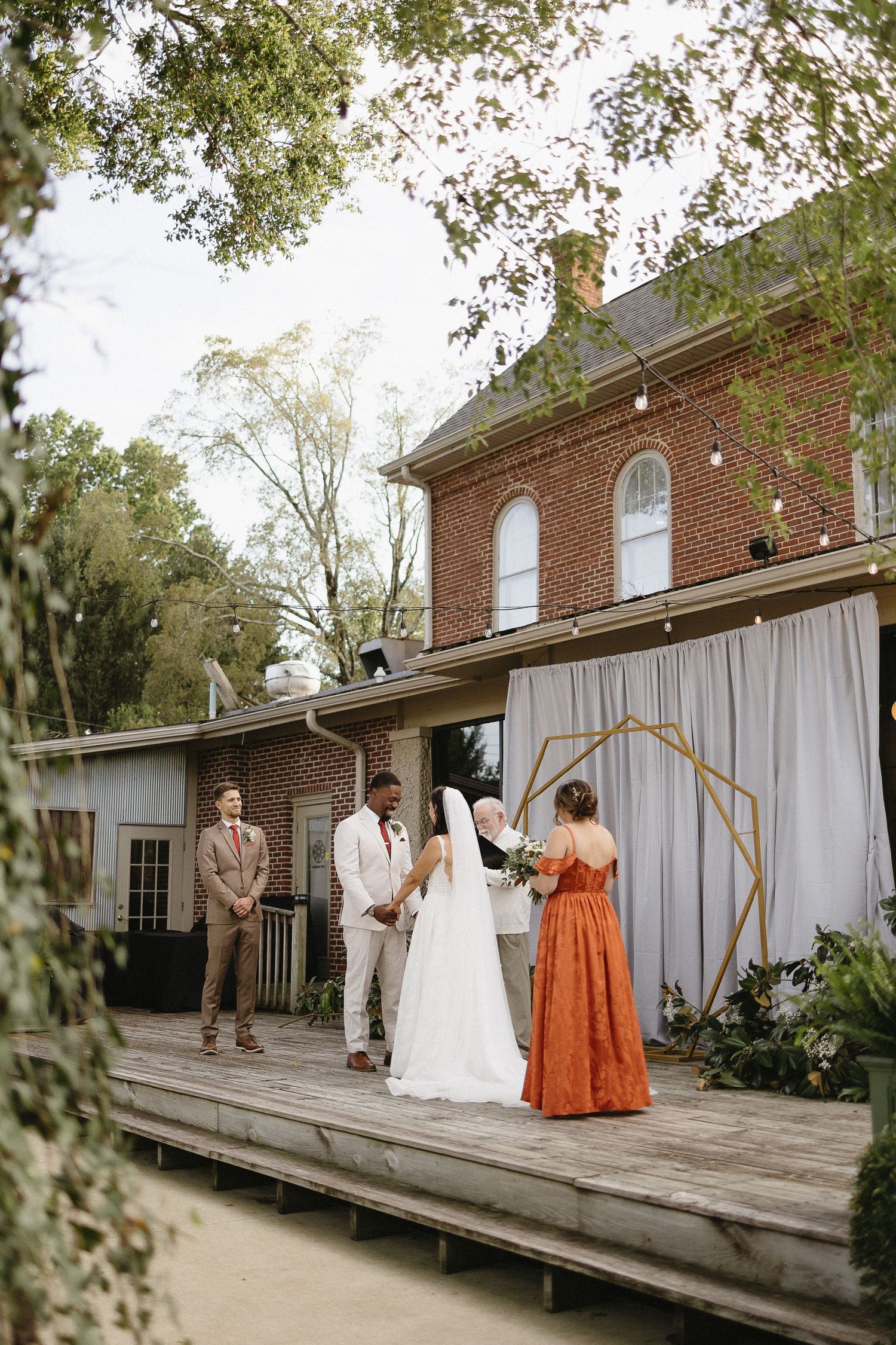 Wedding party poses on wooden deck outside brick venue with white curtain backdrop.