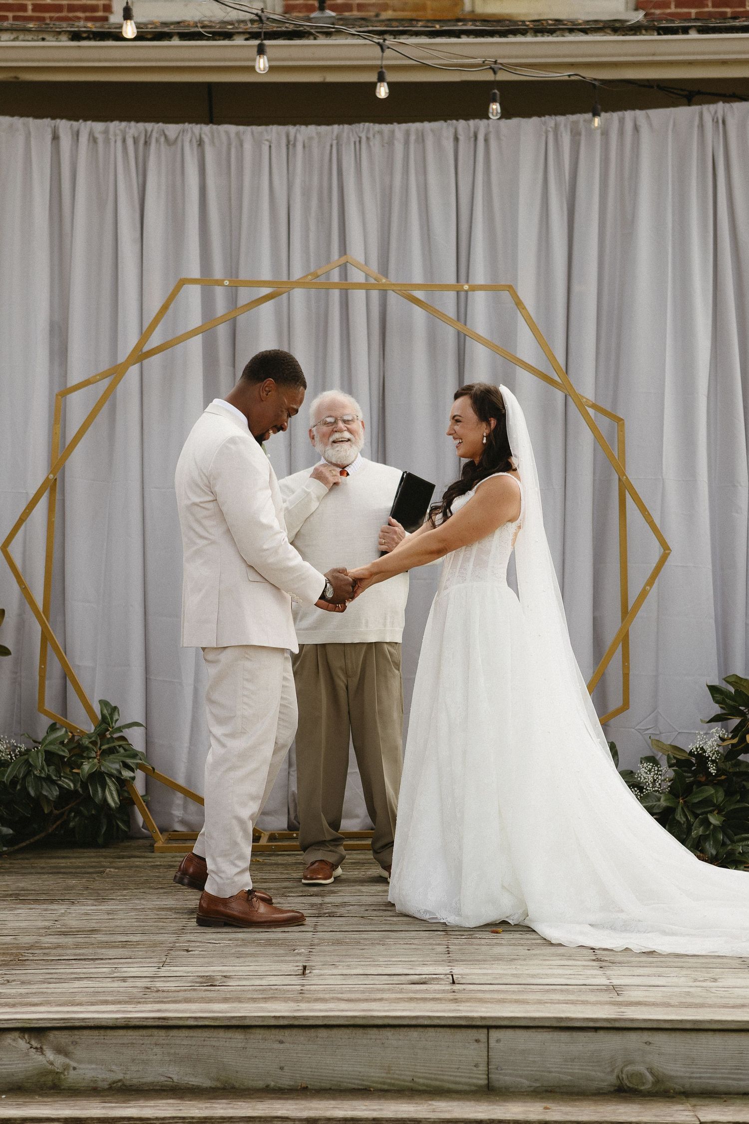 Wedding ceremony taking place under a geometric arch with white curtain backdrop.
