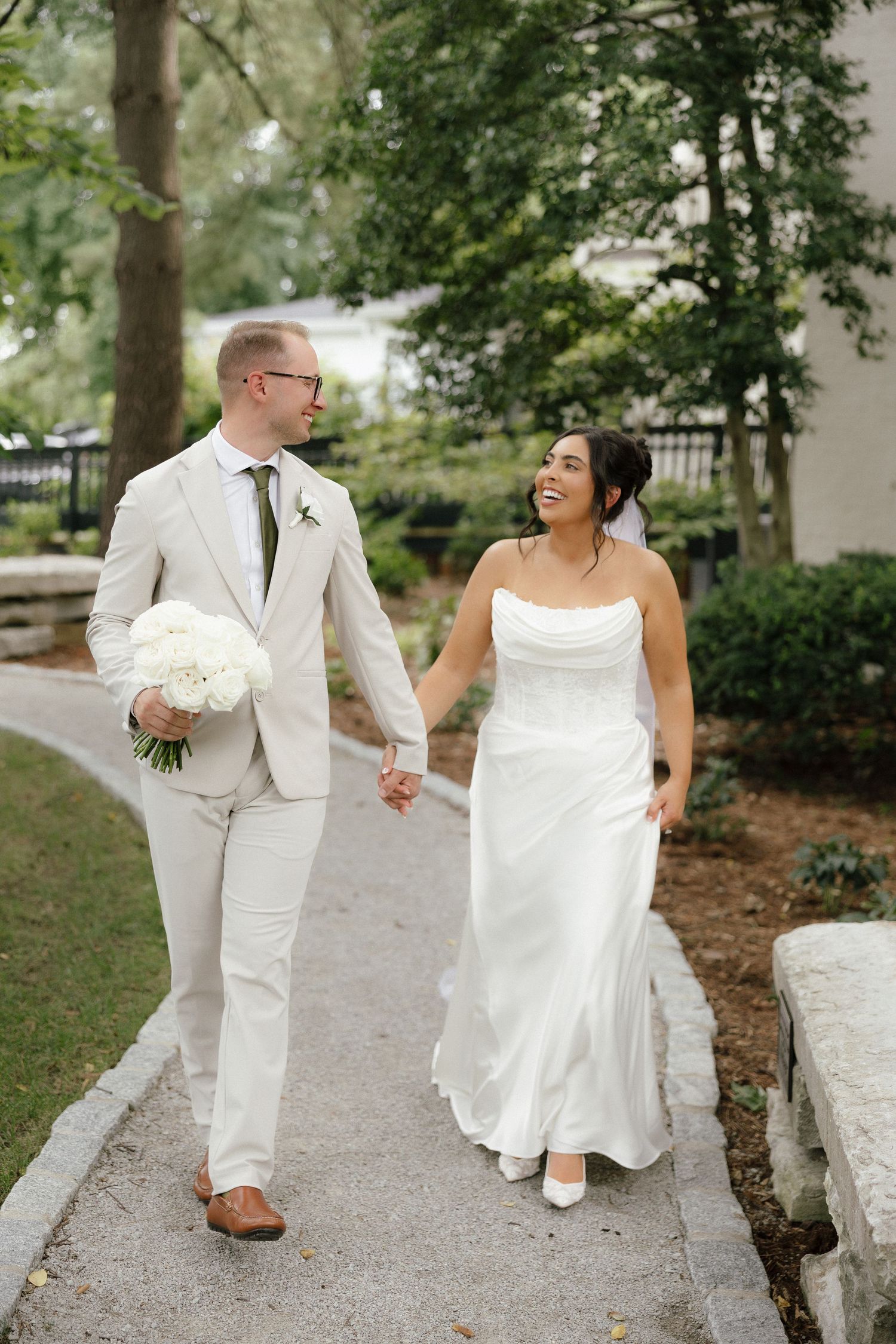 Bride and groom hold hands walking on garden path, groom in tan suit holding white flower bouquet.