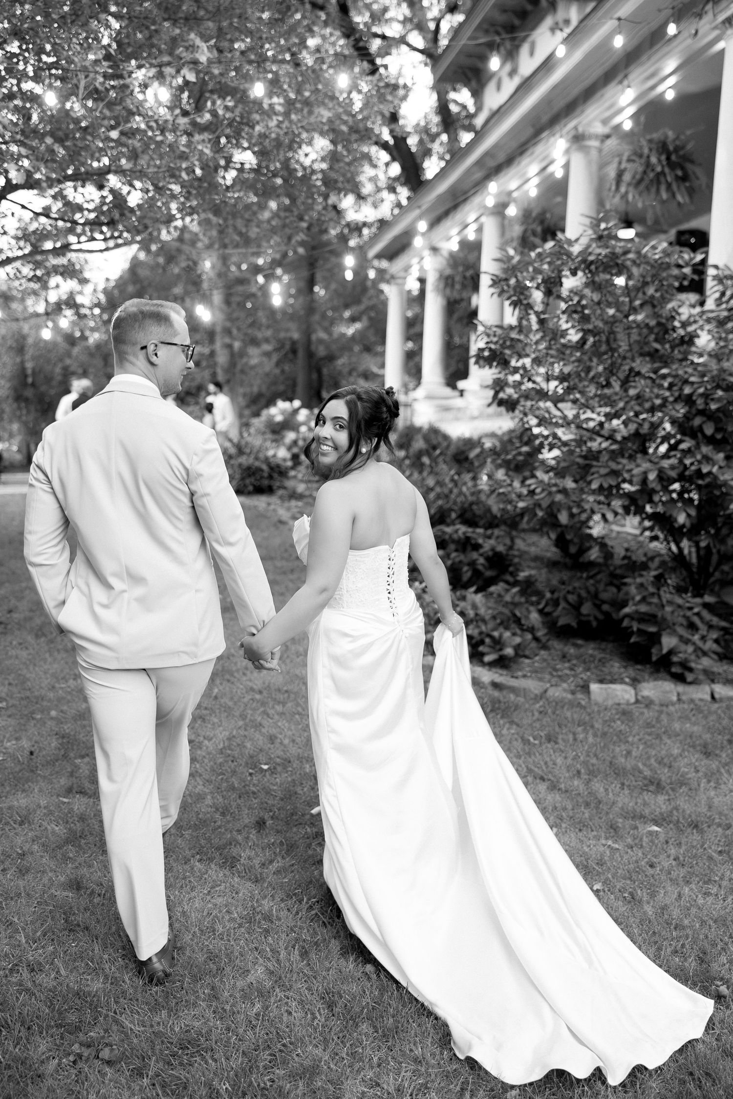 Newlywed couple holds hands walking toward elegant venue with string lights in black and white photo.