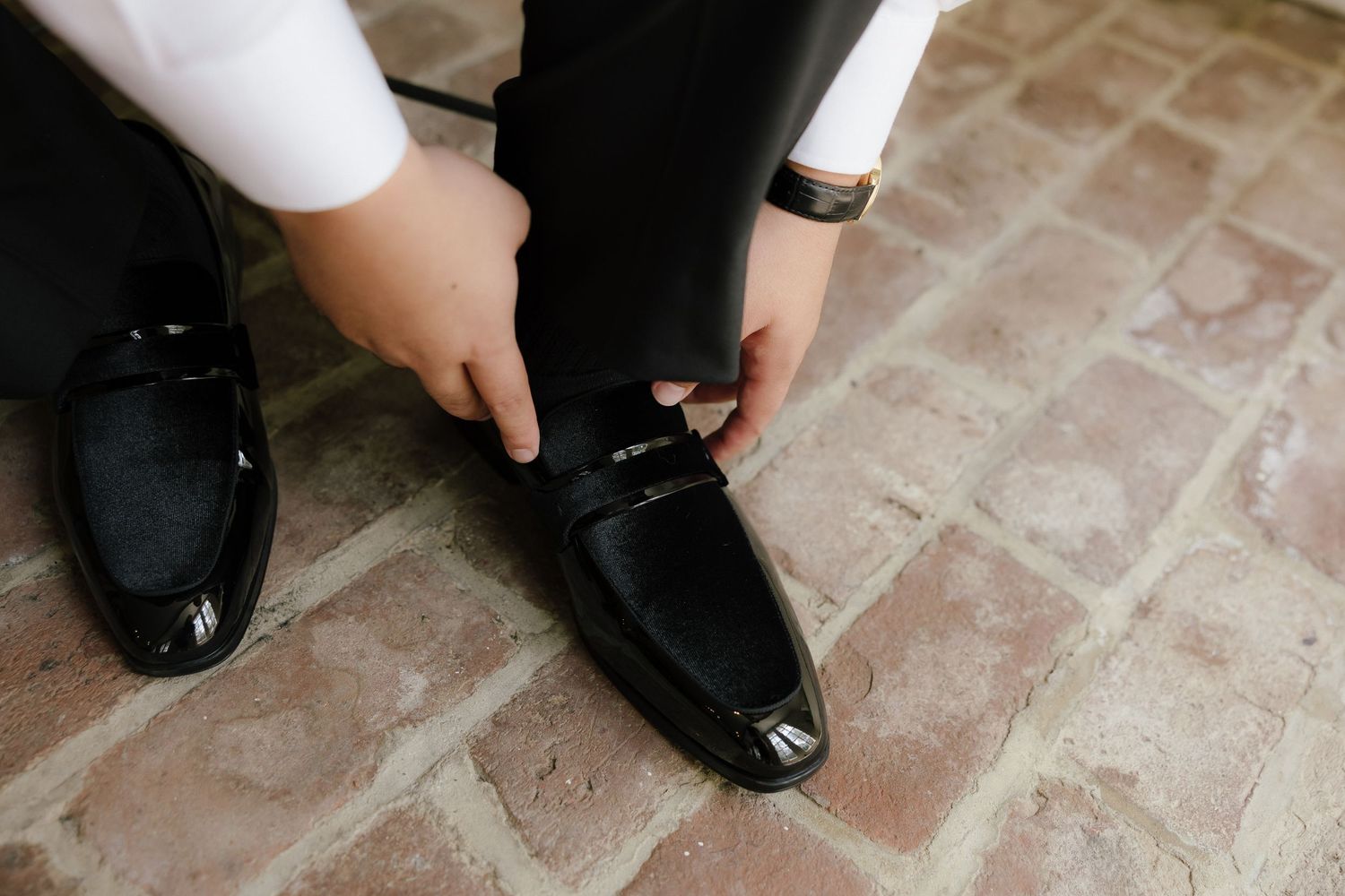 Man putting on shiny black patent leather loafers on brick floor, wearing white dress shirt and dark trousers.