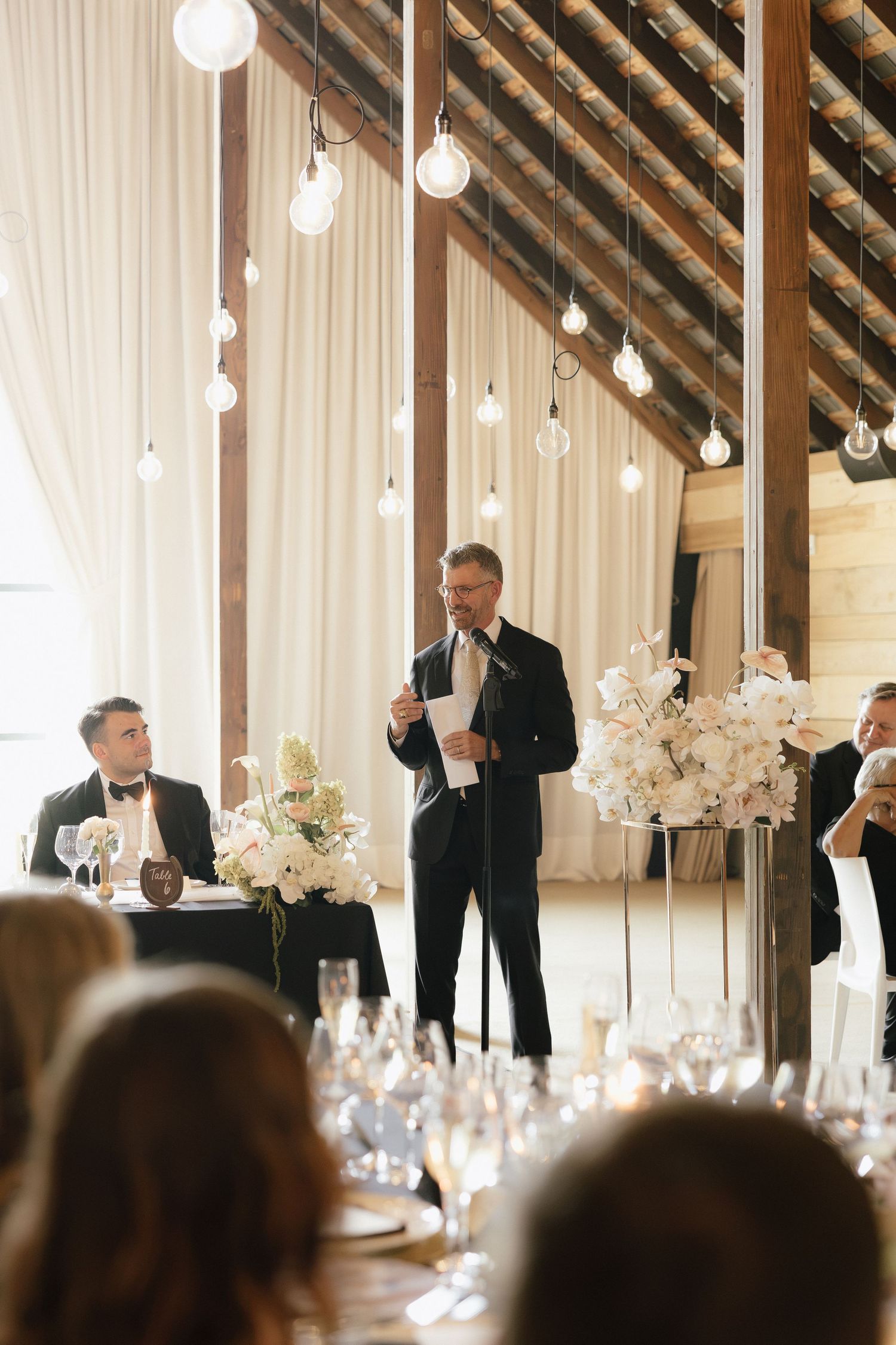 Elegantly dressed speaker delivers toast at rustic barn wedding reception with hanging globe lights and floral arrangements.