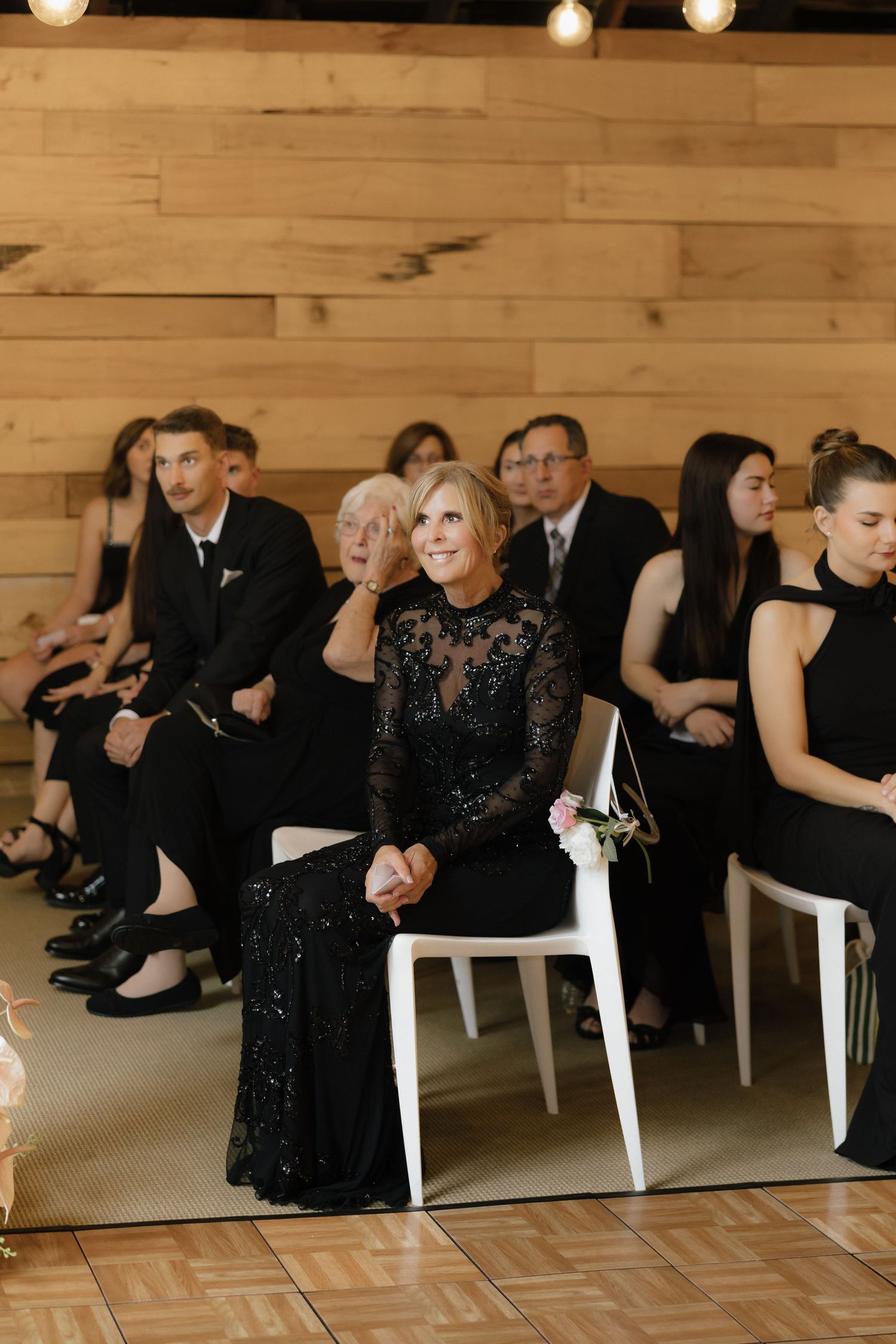 Wedding guests in formal black attire seated at indoor ceremony with warm string lights overhead.